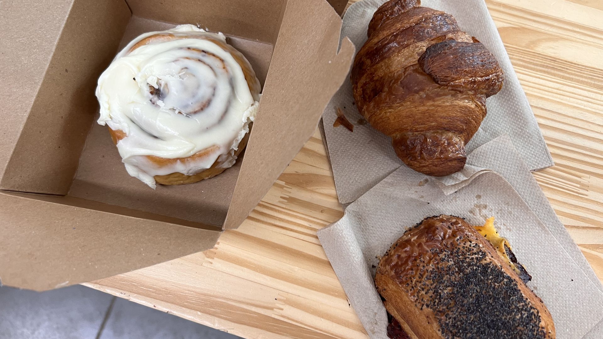 A cinnamon roll with white icing in a brown takeout box sits beside flaky croissants on a napkin, all on a light wooden table.