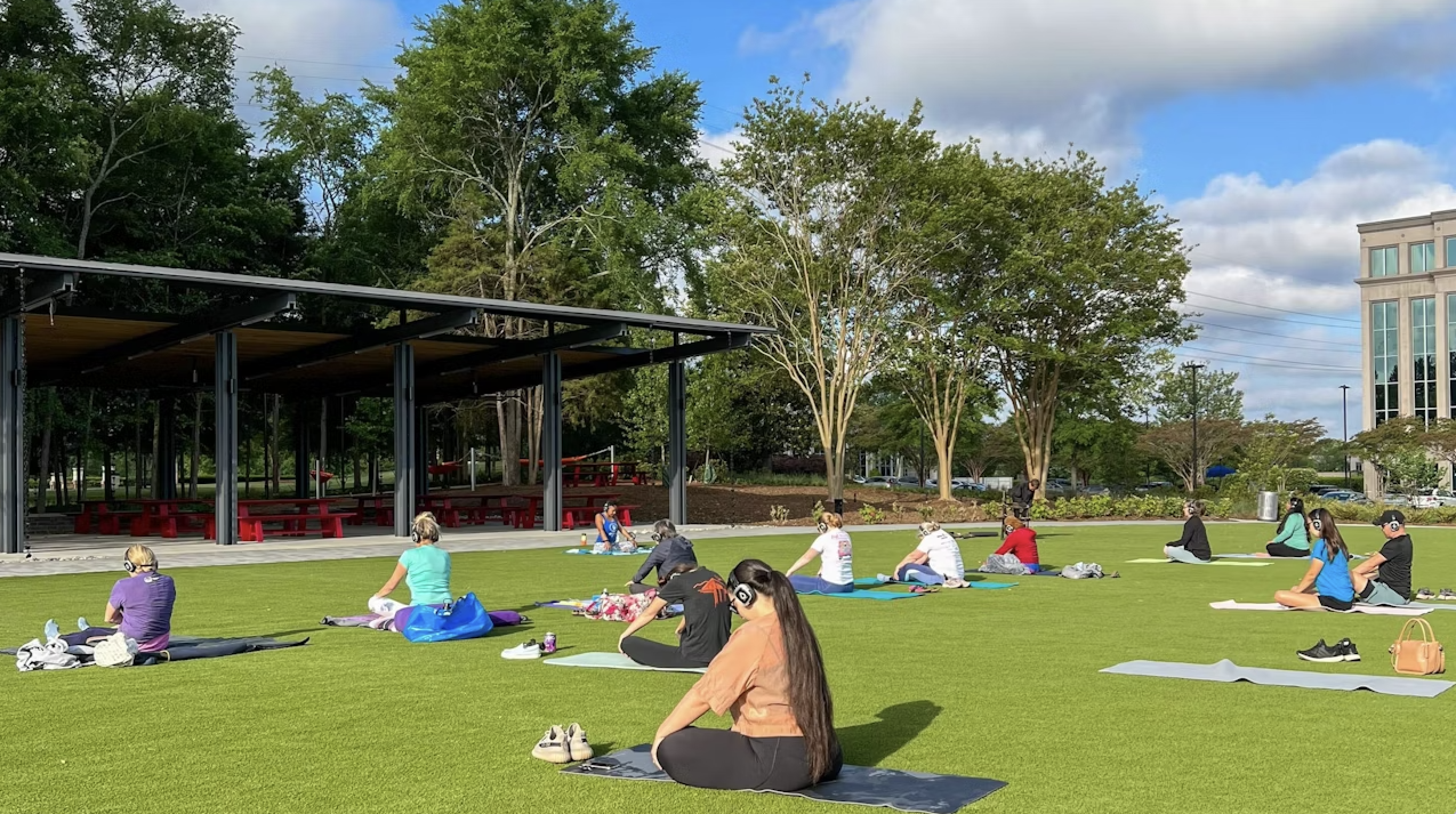 Group of people sitting on yoga mats spaced out on green grass, wearing headphones, under blue sky with white clouds near trees and a pavilion with red benches.