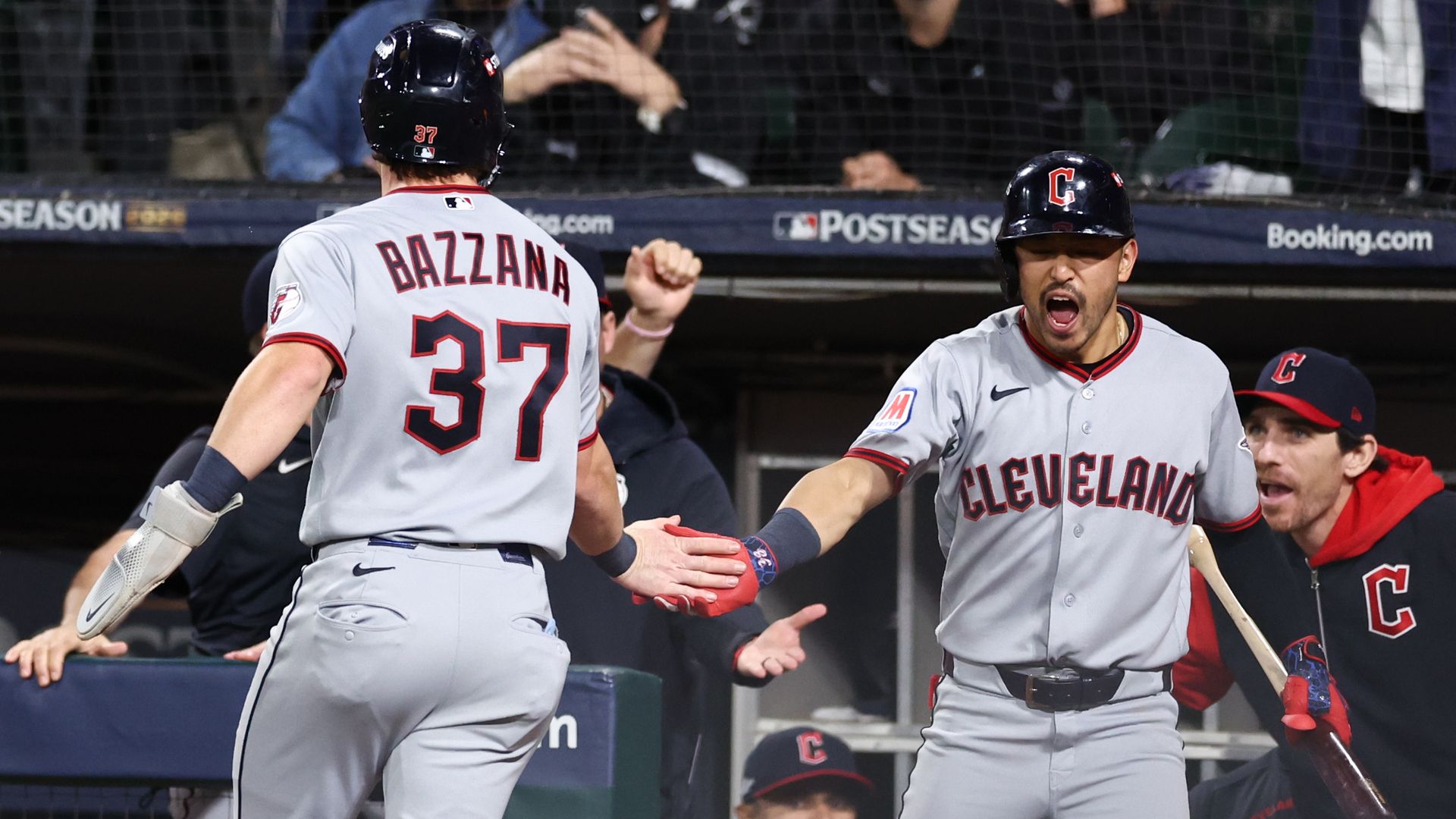 Two Cleveland players in gray uniforms celebrate in the dugout; one with back of jersey reading BAZZANA 37, the other facing forward with helmet and a C logo; fans behind protective netting.