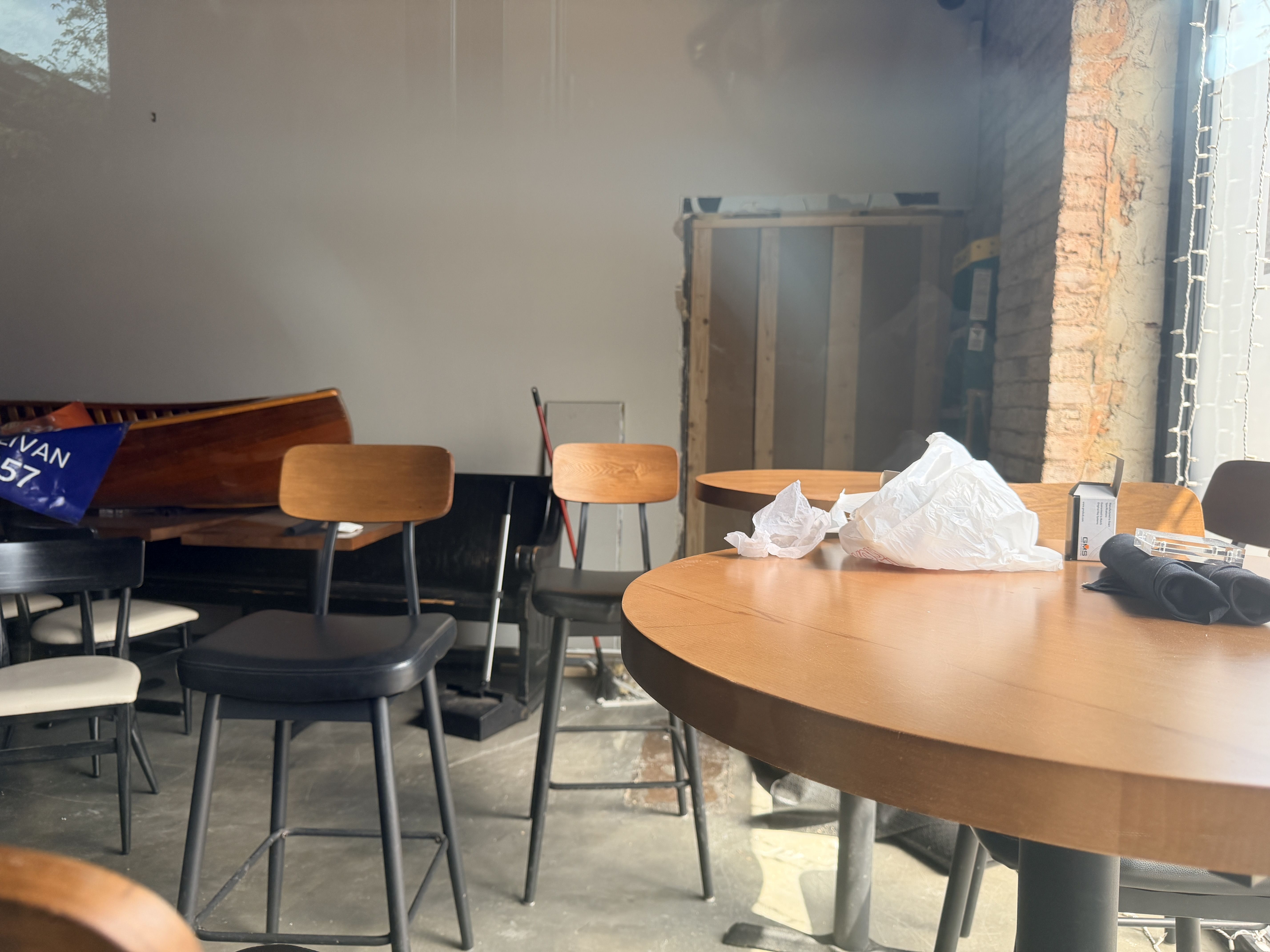 A hazy classroom or cafe with round wooden tables and dark metal-legged chairs. A brick wall with string lights on the right; a wooden bench on the left with a blue banner, and crumpled papers on a foreground table.