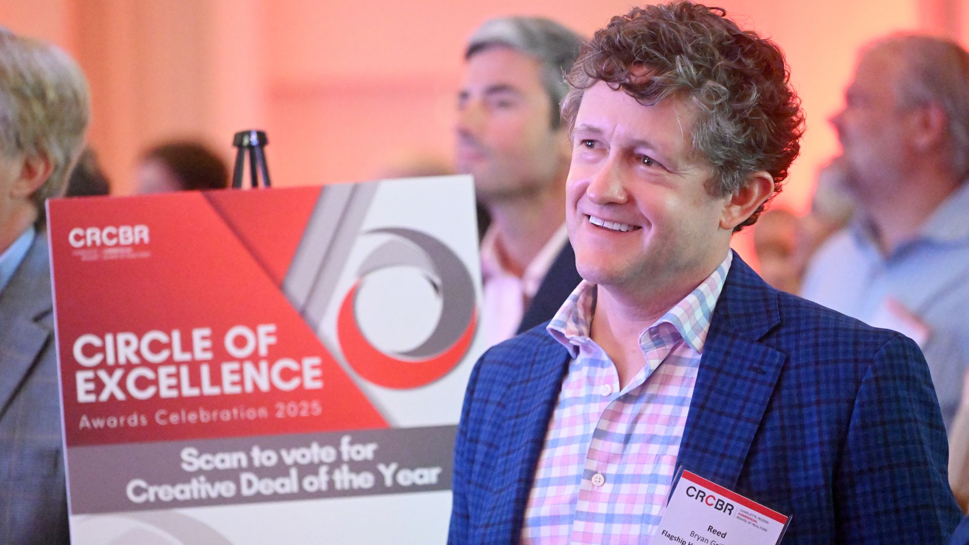 A smiling attendee stands next to a sign for the CRCBR Circle of Excellence Awards Celebration 2025, which invites guests to vote for Creative Deal of the Year.