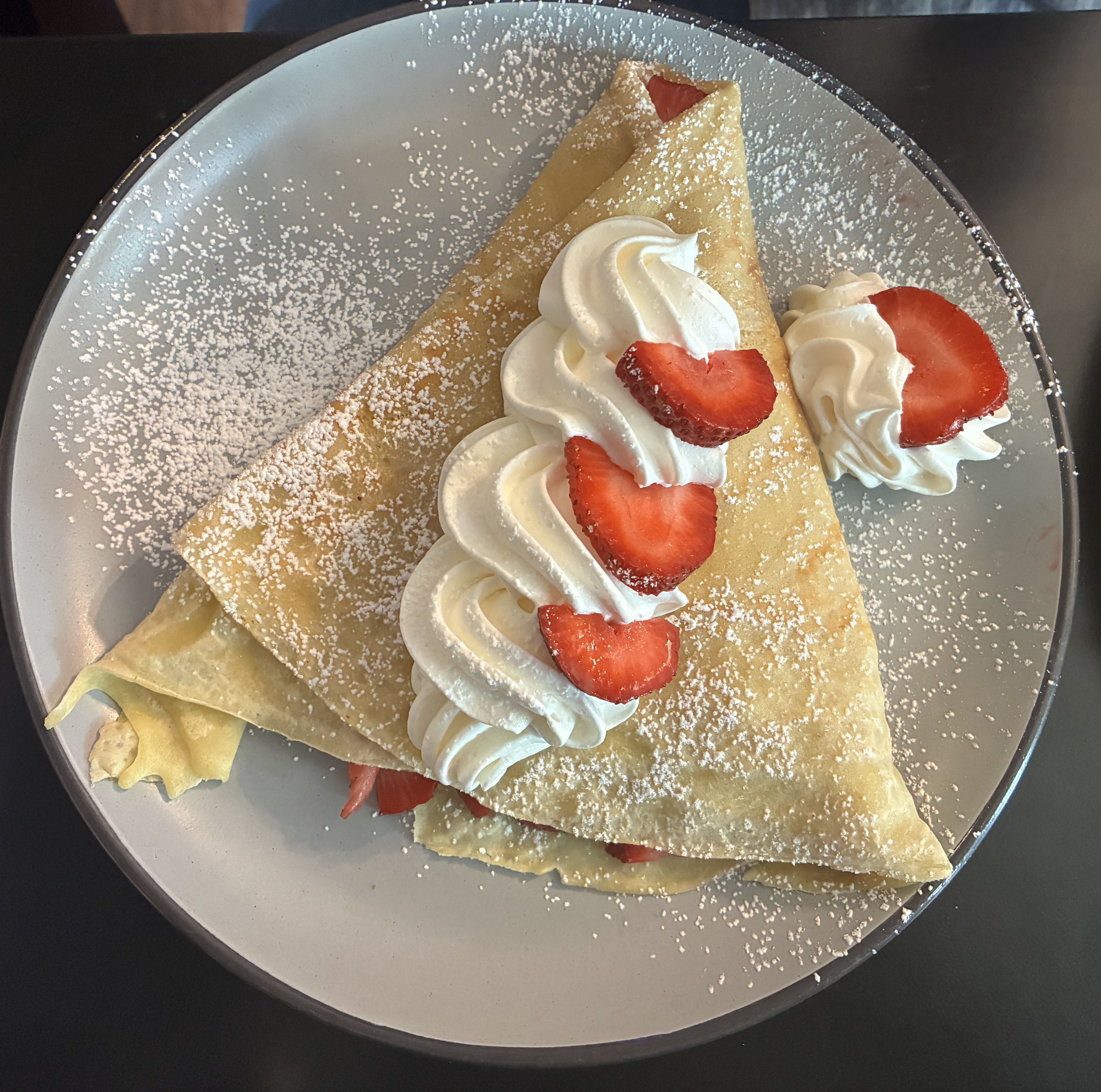 Two folded crepes on a gray plate, topped with swirls of whipped cream and sliced strawberries, lightly dusted with powdered sugar.