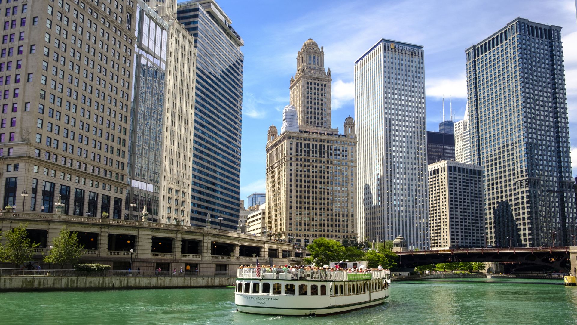 Chicago riverfront skyline featuring beige and glass skyscrapers under a bright blue sky. A white tour boat labeled "Chicago's Leading Lady" sails the emerald-green river in the foreground.
