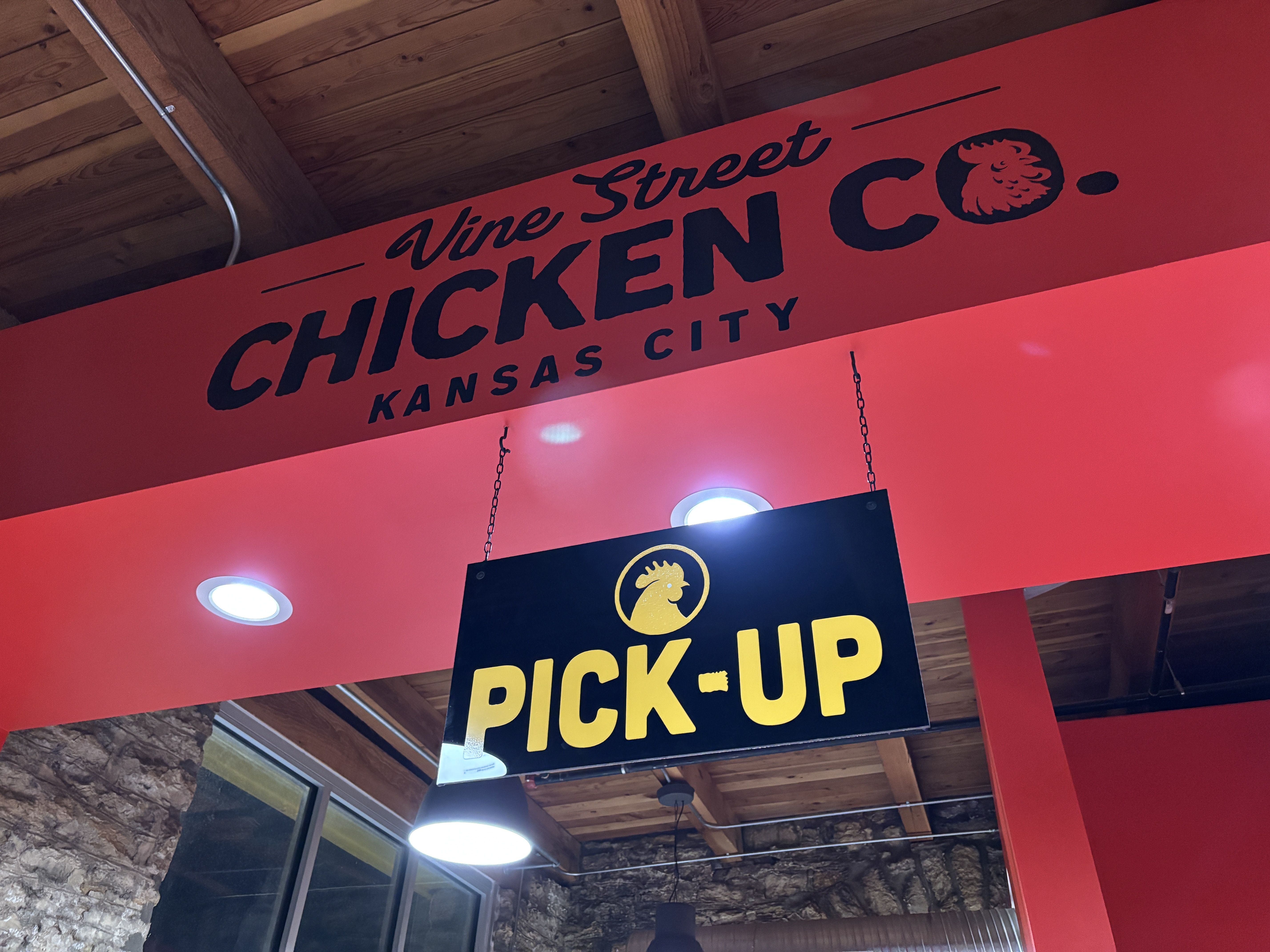 Interior of Vine Street Chicken Co. in Kansas City with red walls and ceiling beams. A black and yellow sign reading "PICK-UP" with a chicken logo hangs from the ceiling.