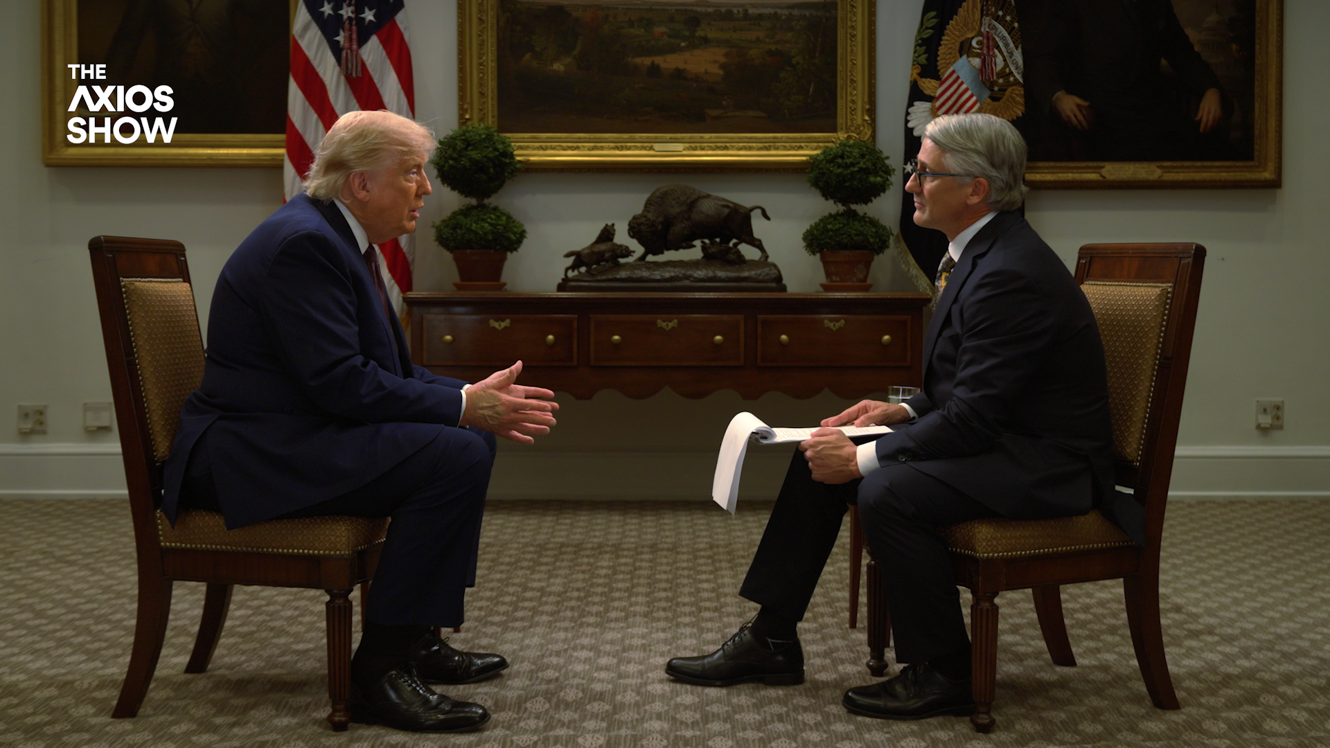 Two men in suits sit facing each other in a formal office. Left resembles Donald Trump; right, gray-haired with glasses, holds papers. A wooden desk and framed paintings sit behind them; Axios Show logo left.