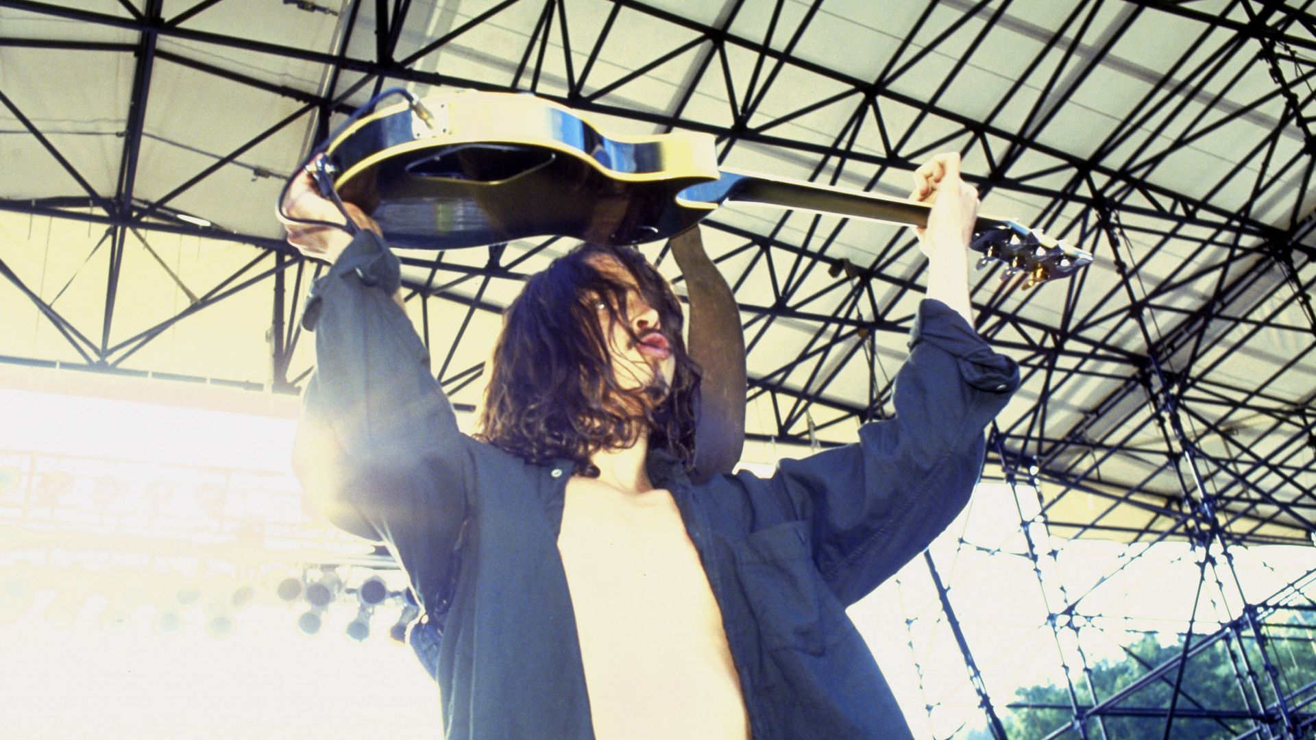 Chris Cornell holds a guitar over his head with a stage roof visible above him. He's wearing a 90sera button up shirt unbuttoned over a T-shirt, his hair is long.