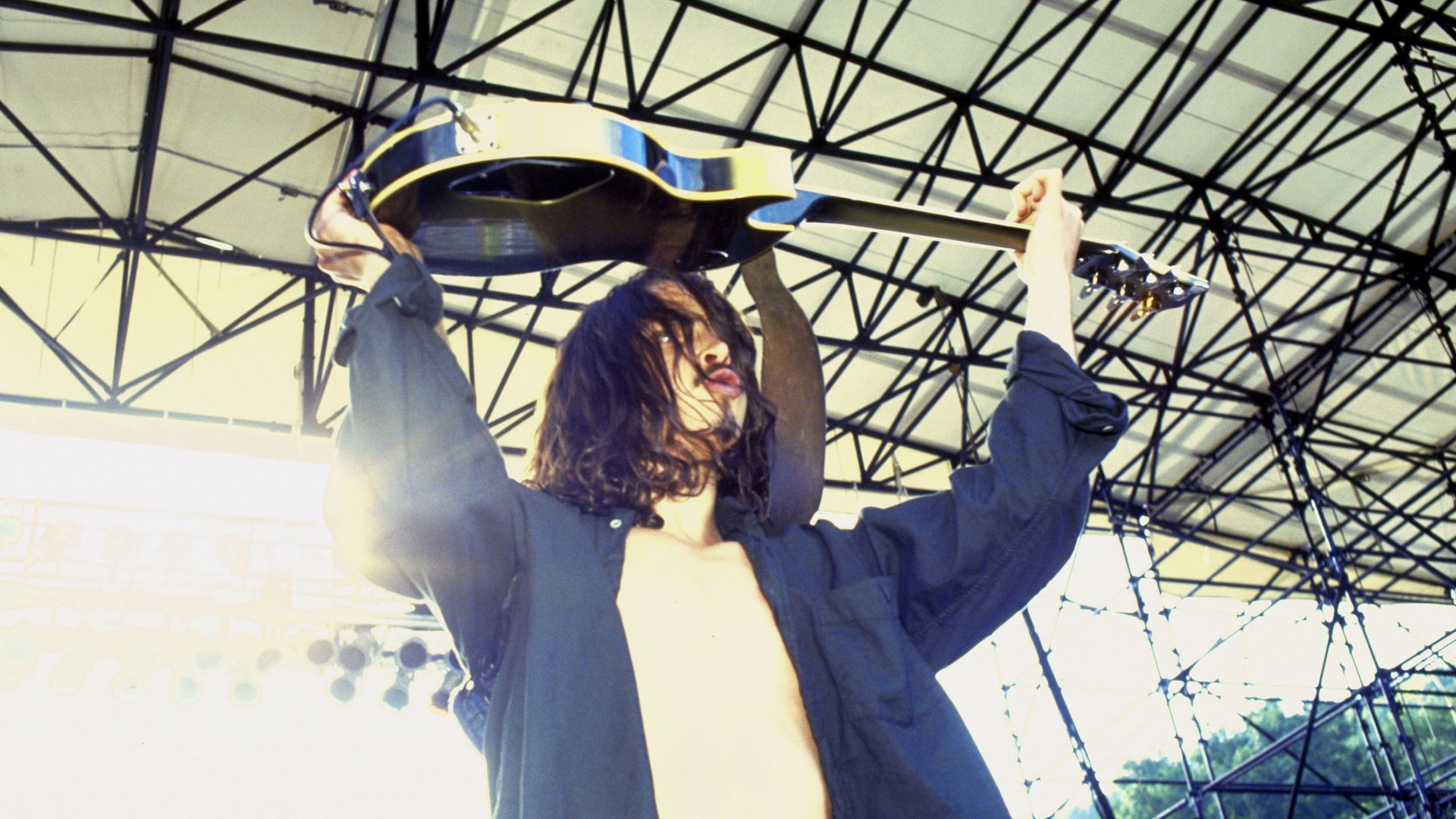 Chris Cornell holds a guitar over his head with a stage roof visible above him. He's wearing a 90sera button up shirt unbuttoned over a T-shirt, his hair is long.