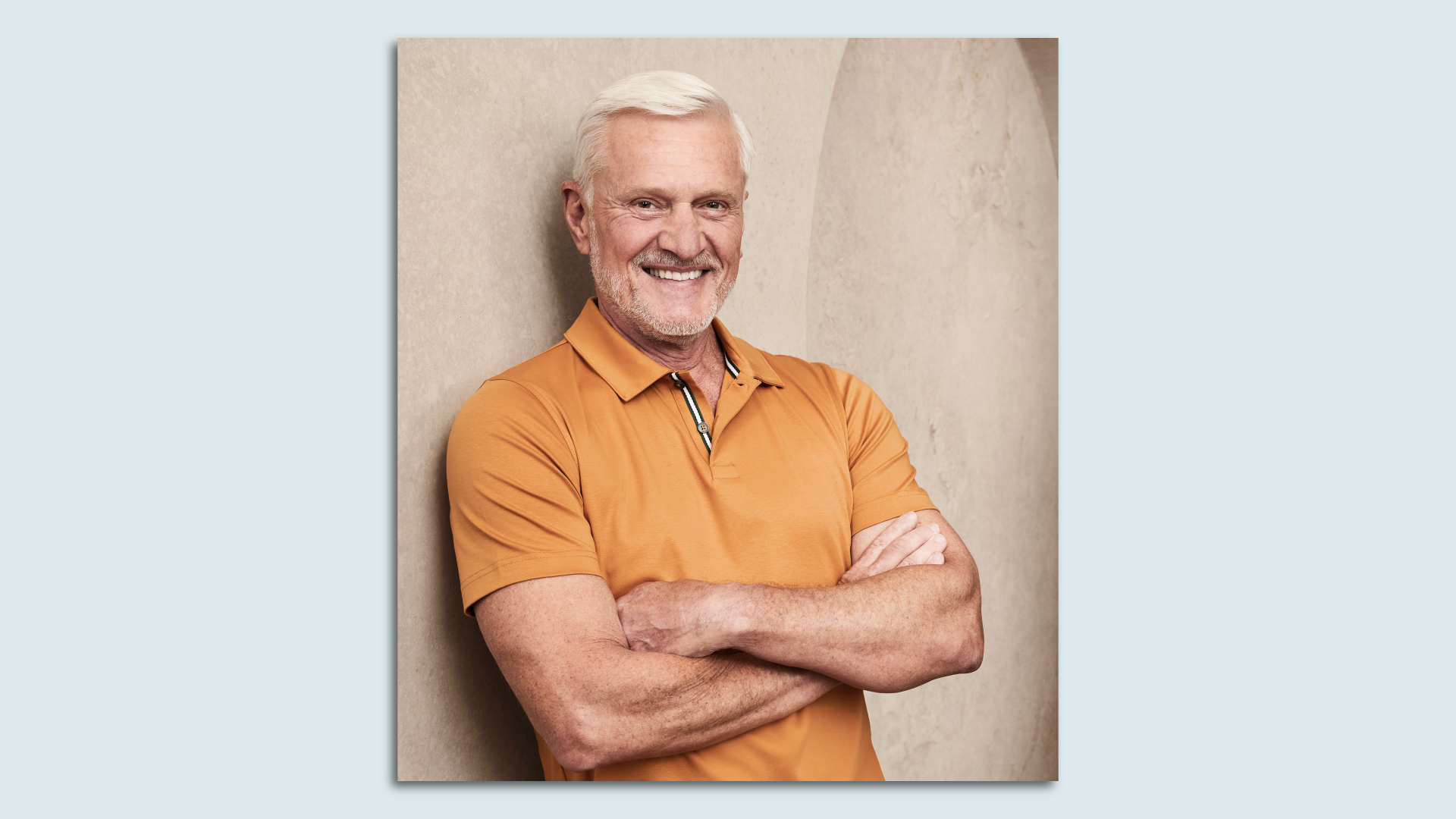 A man with white hair and a beard crosses his arms and smiles, leaning against a wall in an orange polo shirt.
