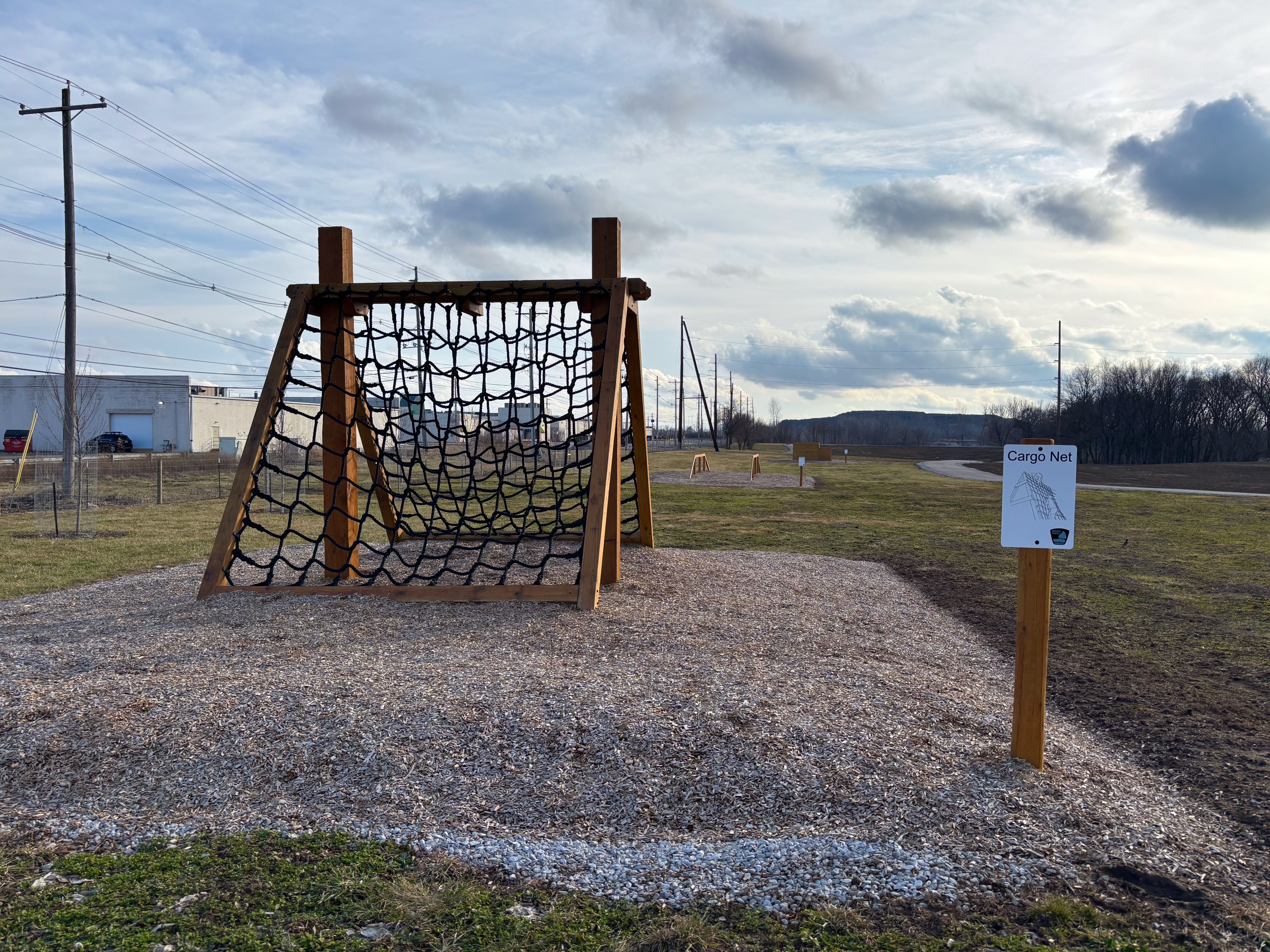 Outdoor fitness area with a wooden cargo net climbing structure on wood chips, a sign labeled "Cargo Net," grass field, and overcast sky with scattered clouds.