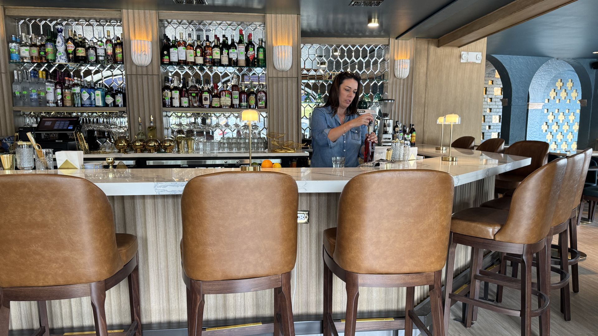 Woman in denim shirt opening a bottle behind a marble-top bar with brown leather stools and shelves of liquor bottles and glasses in a modern bar interior.