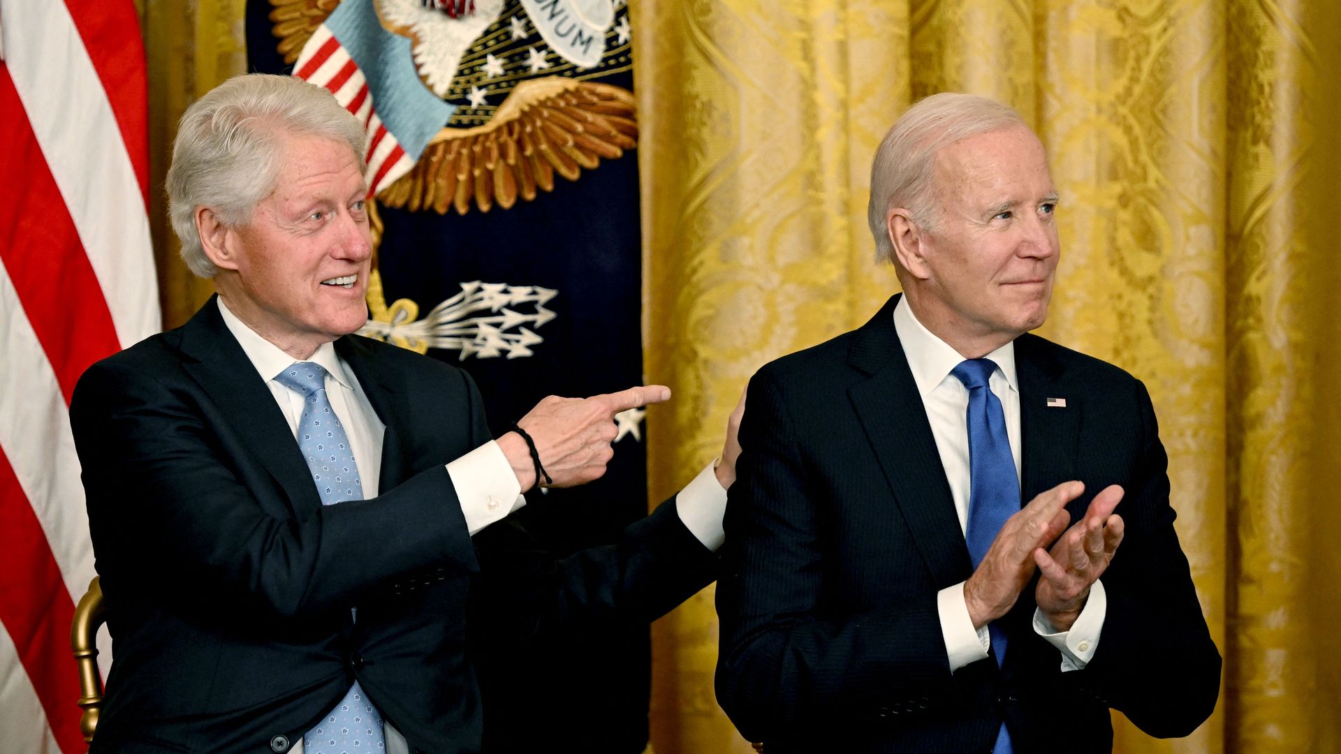 Former Presidents Clinton and Biden, both with gray-white hair and both in black jackets, white shirts and blue ties, gesture during an event in front of a US flag and gold curtain at the White House.