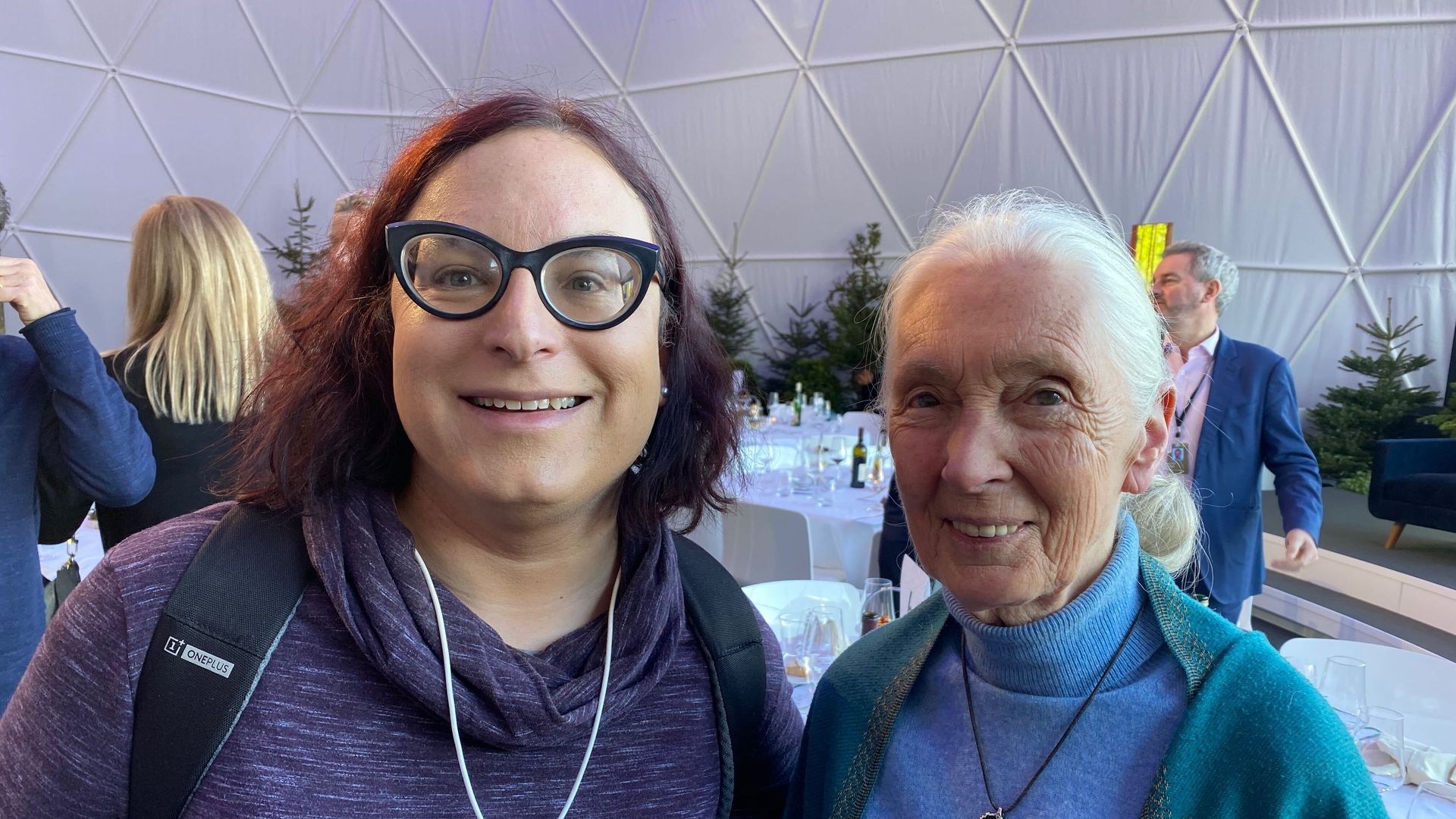 Two smiling women indoors at an event with white dome walls and tables behind. One woman wears large black glasses, a purple top with a press badge, the other a blue turtleneck and shawl with Africa pendant. Also, one is Jane Goodall and the other is Axios Chief Technology Correspondent Ina Fried.