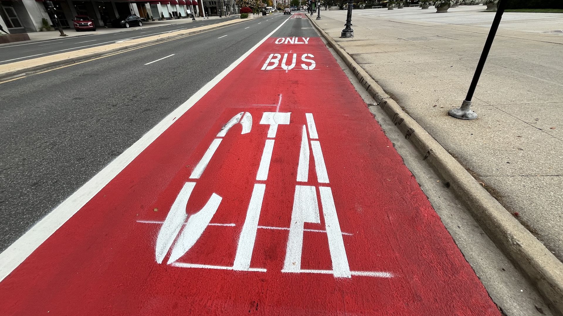 Urban city street with a bold red bus-only lane marked with "ONLY BUS" running down the center; left side has buildings, while a wide sidewalk with planters sits on the right.