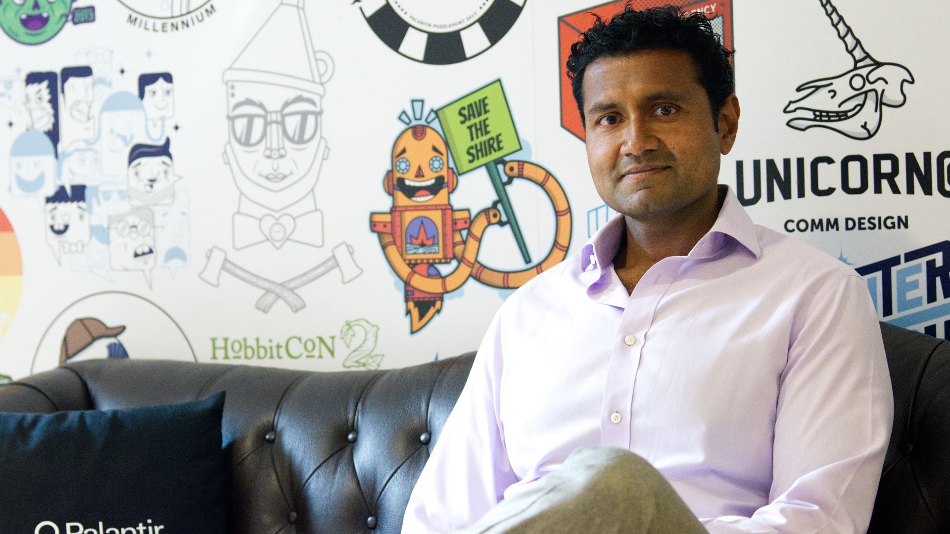 Shyam Sankar, a man, sits on a leather couch. He is to the right of the frame. A Palantir pillow is to the left. Behind him, a white wall with colorful company and event logos.