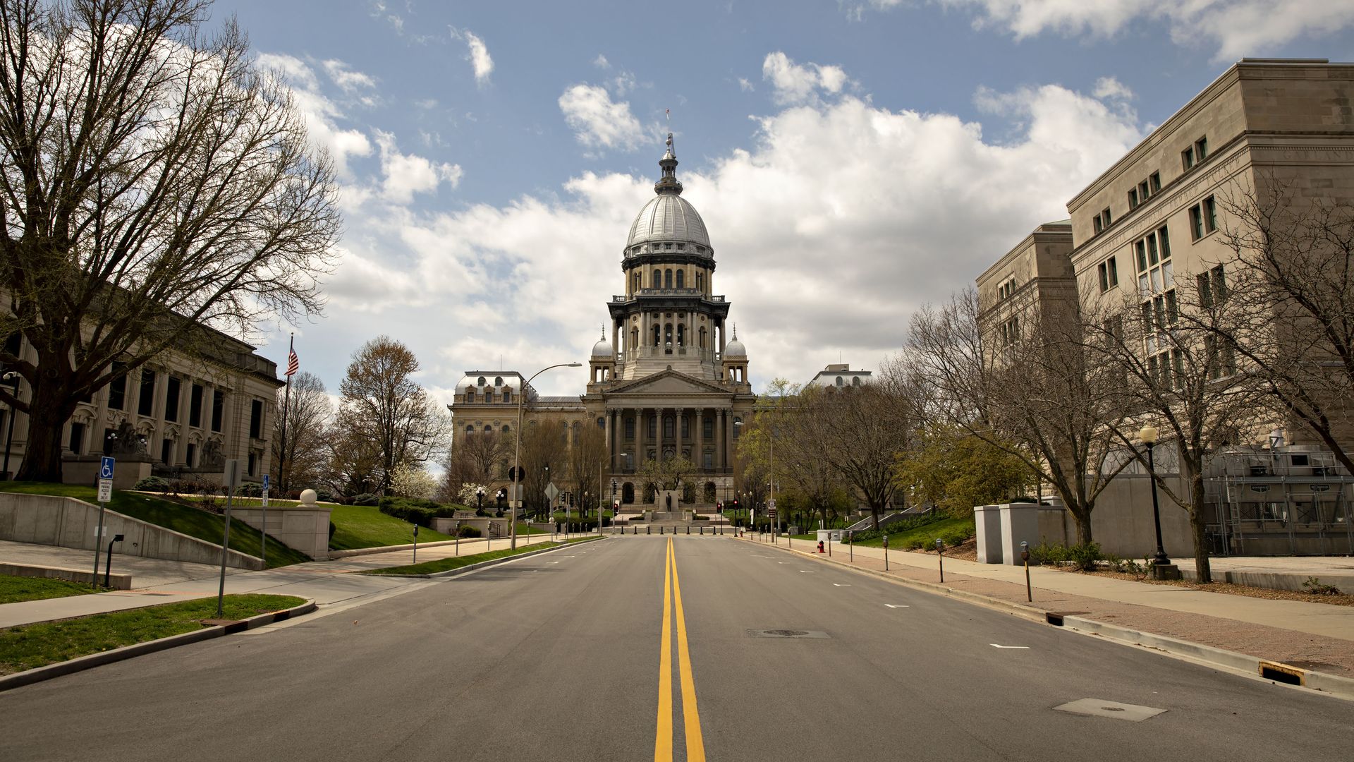 Wide avenue leads to a domed capitol-style government building with columns, flanked by leafless trees and beige side buildings; a double-yellow line runs toward the structure under a cloudy blue sky.