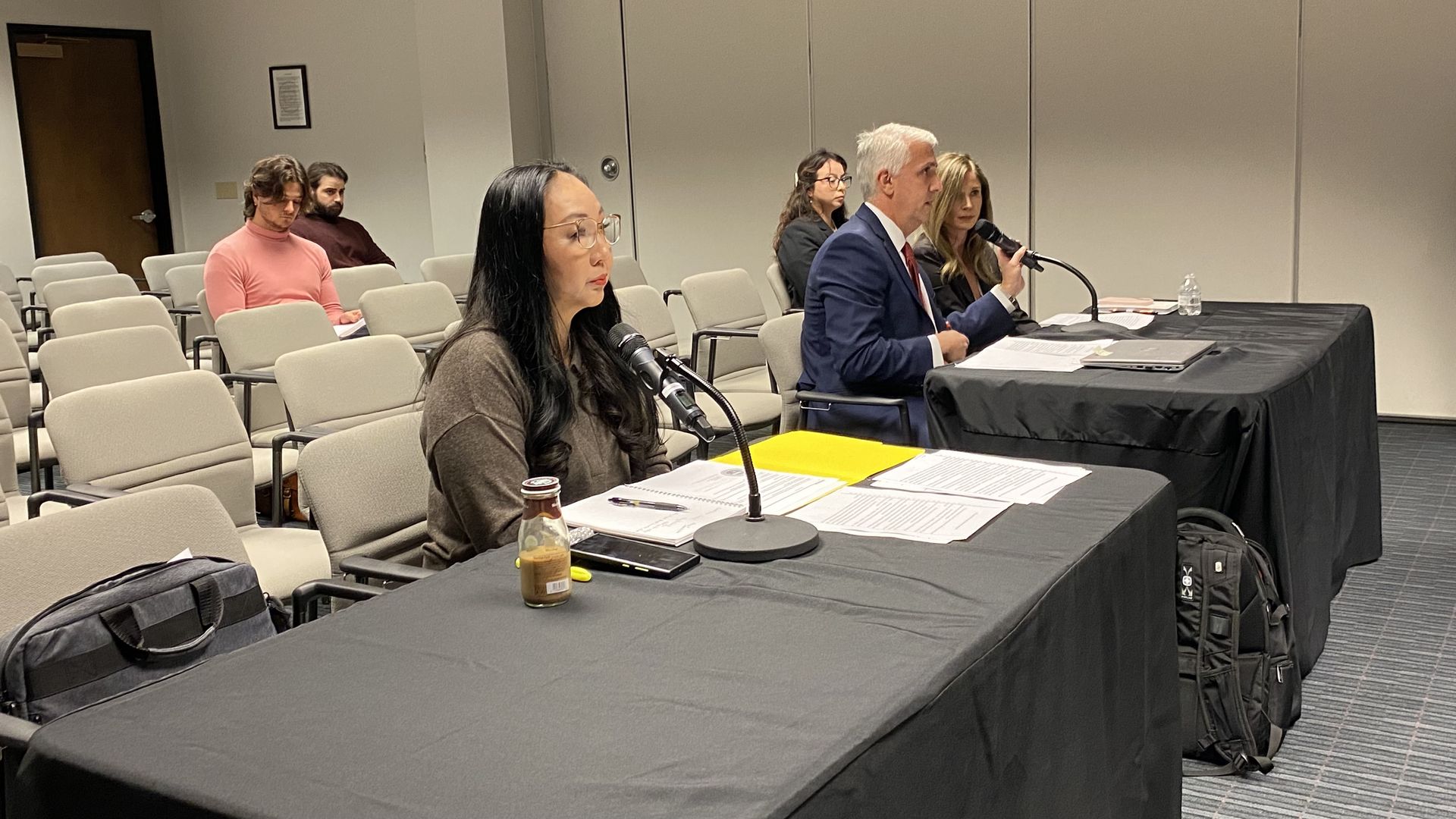 A woman seated at a table with a microphone with two people sitting at an adjacent table.