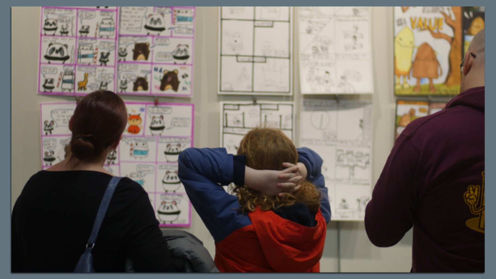 Three people viewing colorful comic strips and drawings displayed on a wall, one in black with a blue strap bag, one in a blue and red jacket with hands behind head, and one in a burgundy hoodie.