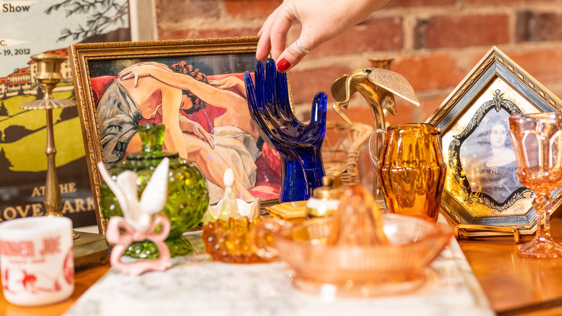 A hand with red nail polish touches a blue glass hand sculpture on a table filled with vintage glassware, a framed painting of embracing figures, and a portrait in a gold frame against a brick wall.
