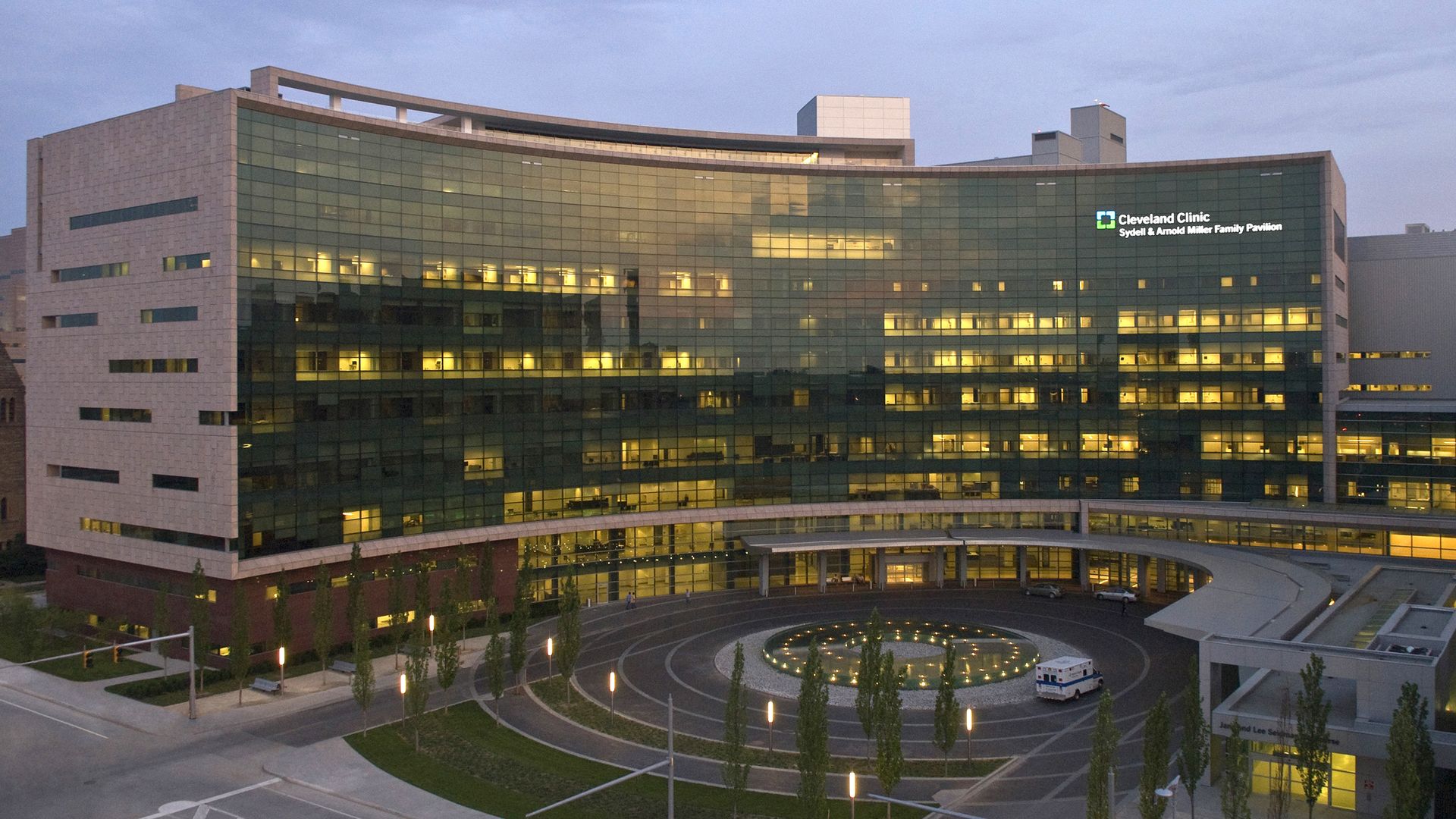 Evening view of Cleveland Clinic Sydell & Arnold Miller Family Pavilion, a modern glass building with yellow interior lights on. Ambulance parked near circular driveway with lights and trees.