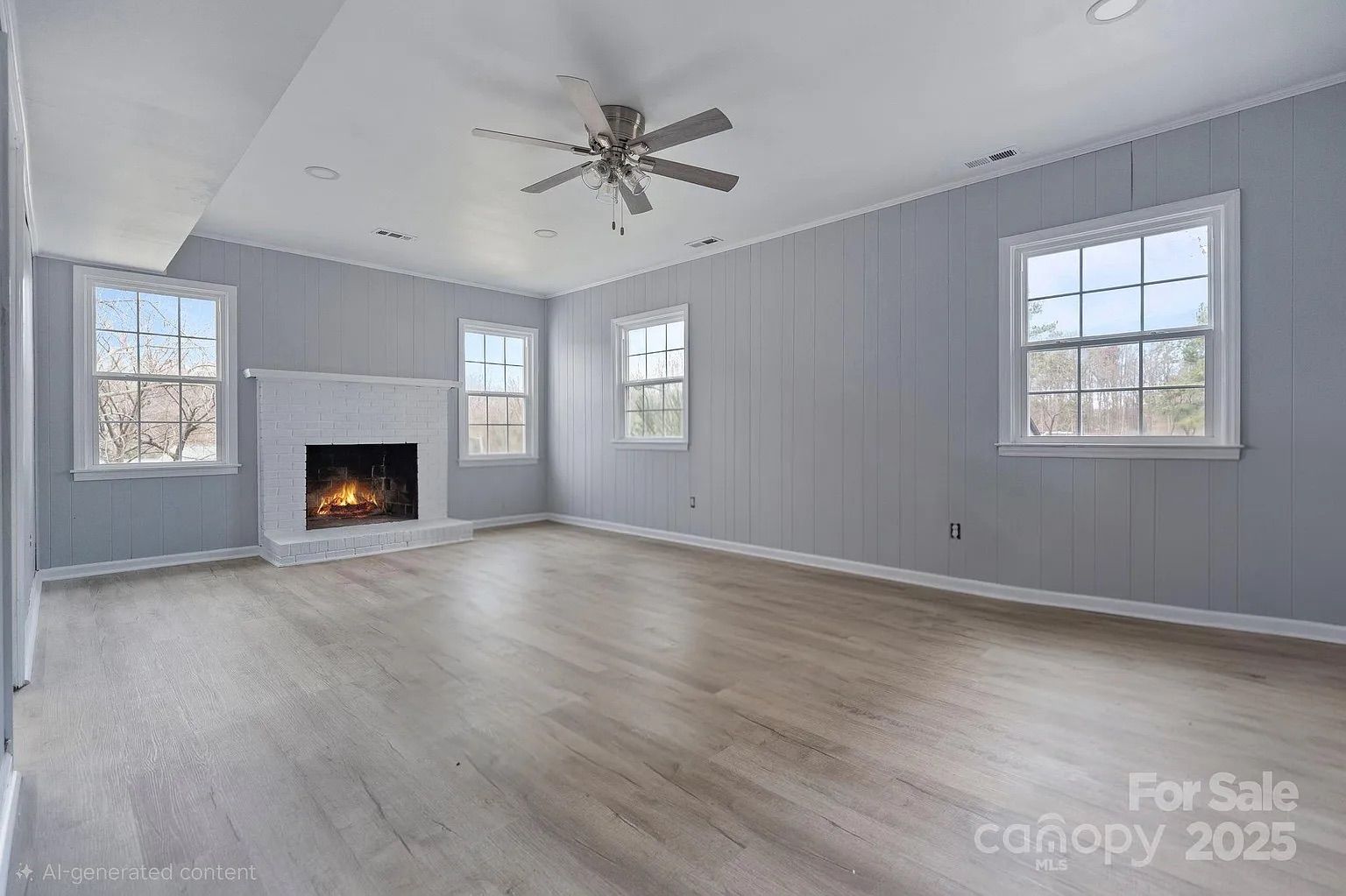 Empty room with light gray panel walls, four white-trimmed windows showing trees outside, a white brick fireplace with a small fire, light wood floor, and ceiling fan with lights.