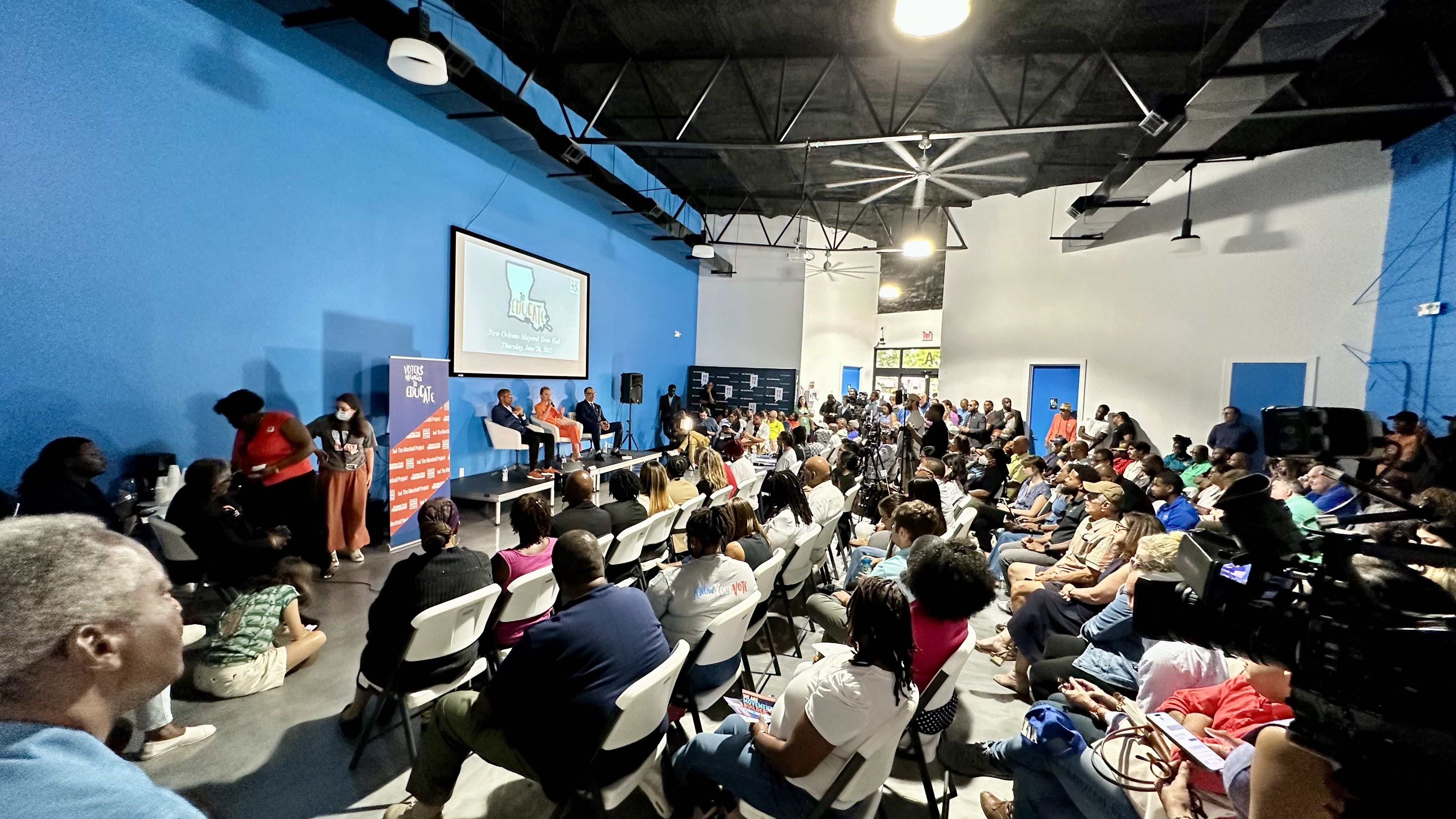 A large group of people attending a panel discussion in a spacious room with blue walls and high ceilings. The audience is seated and facing a stage where the panelists are speaking. A large screen behind the panel displays text related to the event. The room is equipped with professional lighting a