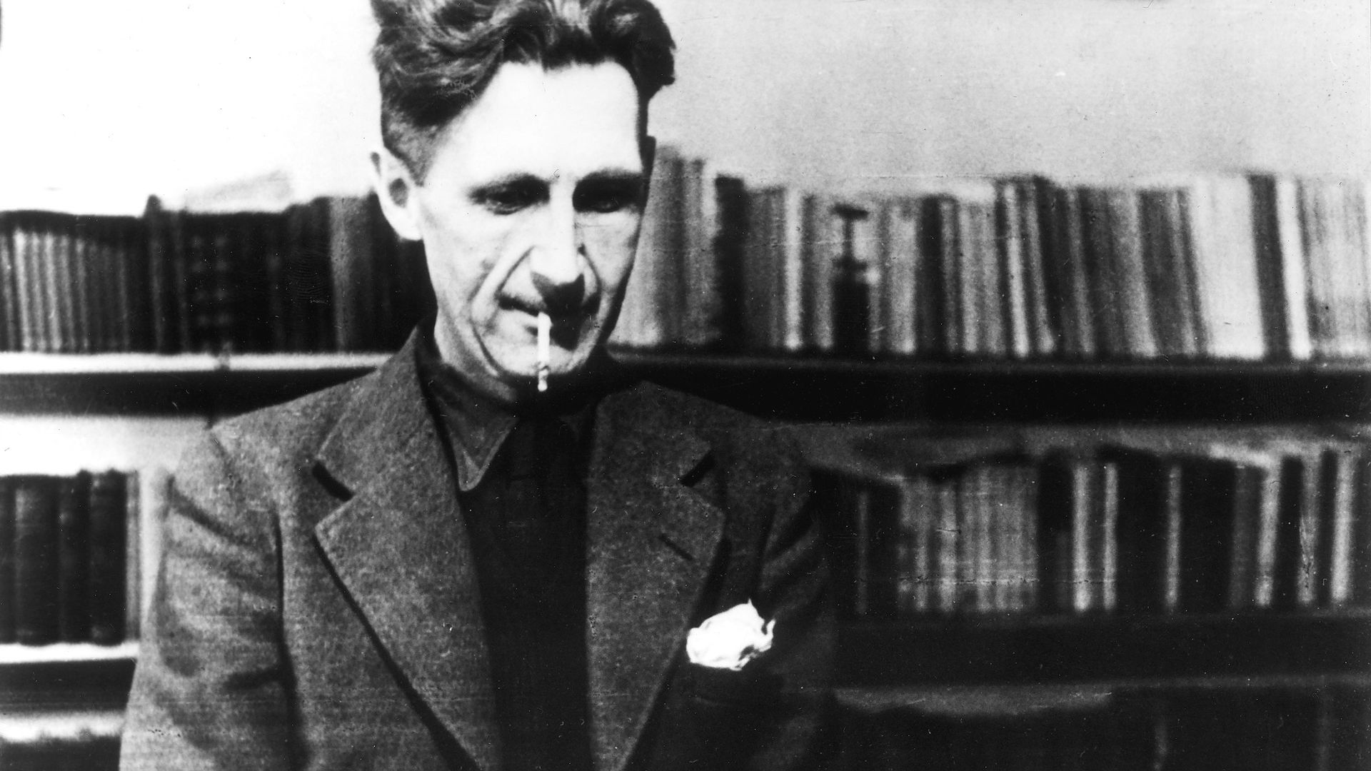 Black and white photo of George Orwell in a suit with a cigarette in his mouth, sitting at a desk typing on a typewriter, with bookshelves filled with books in the background.