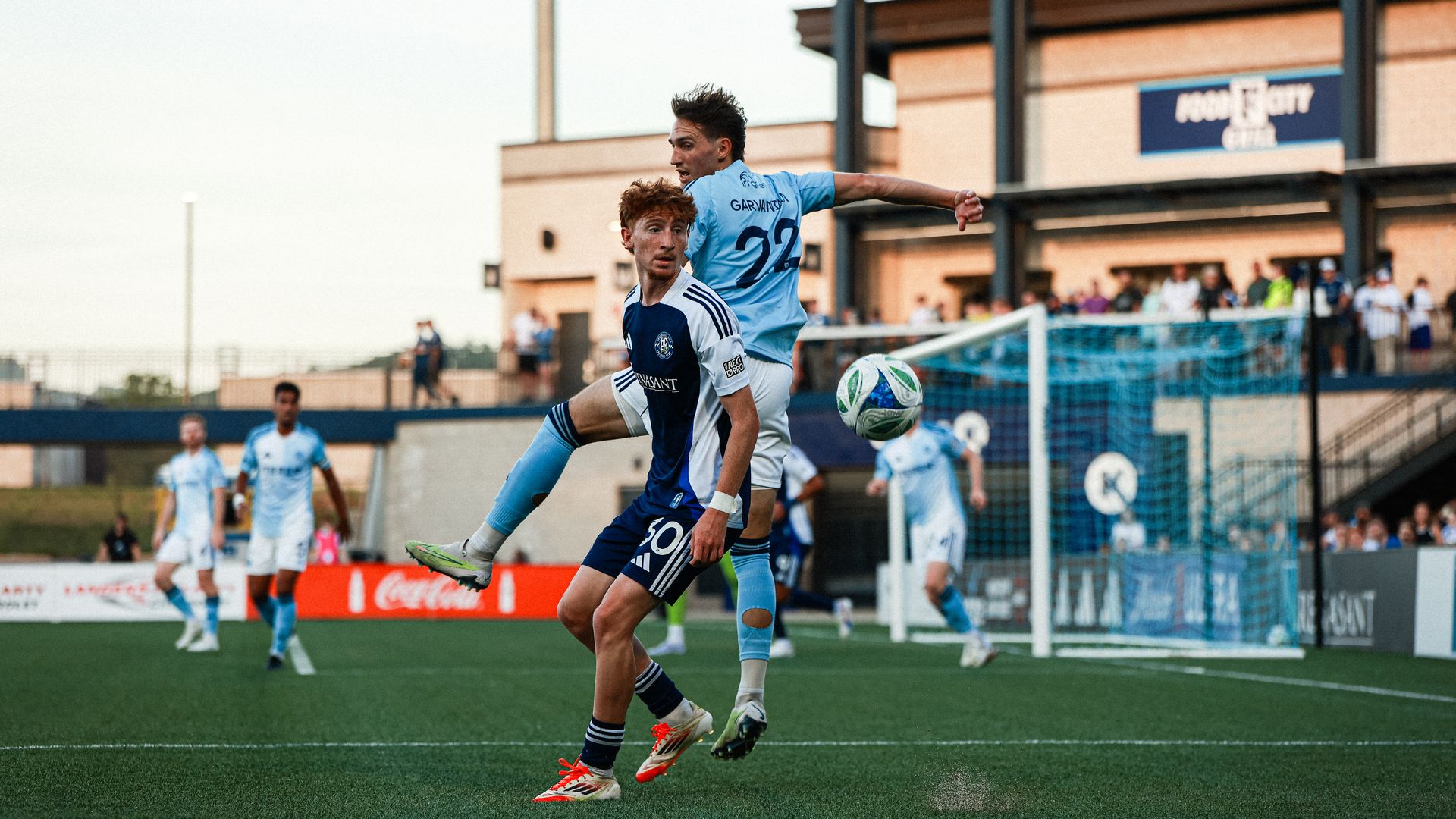 Two soccer players in blue uniforms compete for the ball near the goal at Wicks Family Field, with other players and spectators visible in the background.