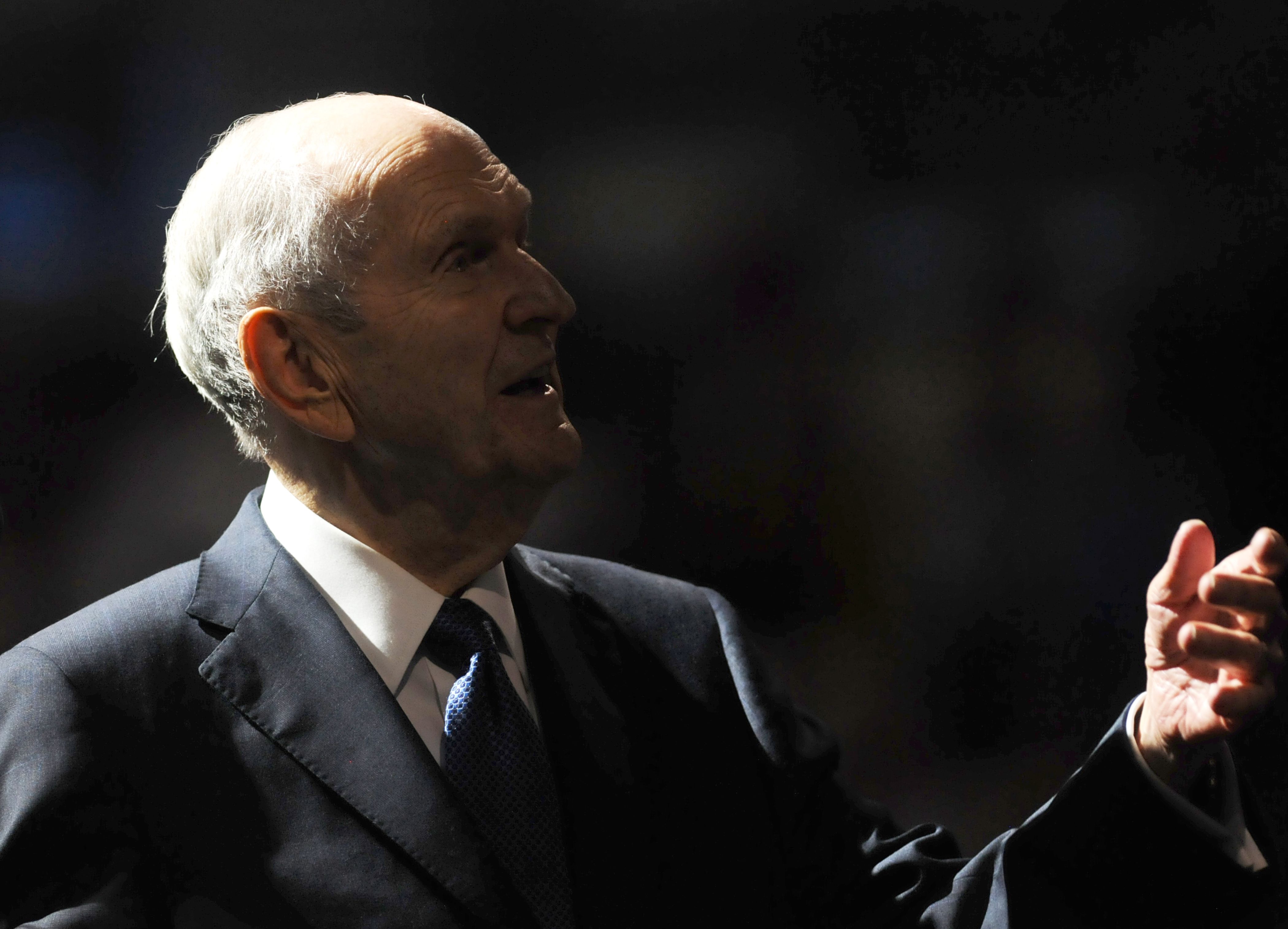 Elderly man in a dark suit and blue tie speaking or gesturing with hand against a dark background, lit from above highlighting his profile and hand.