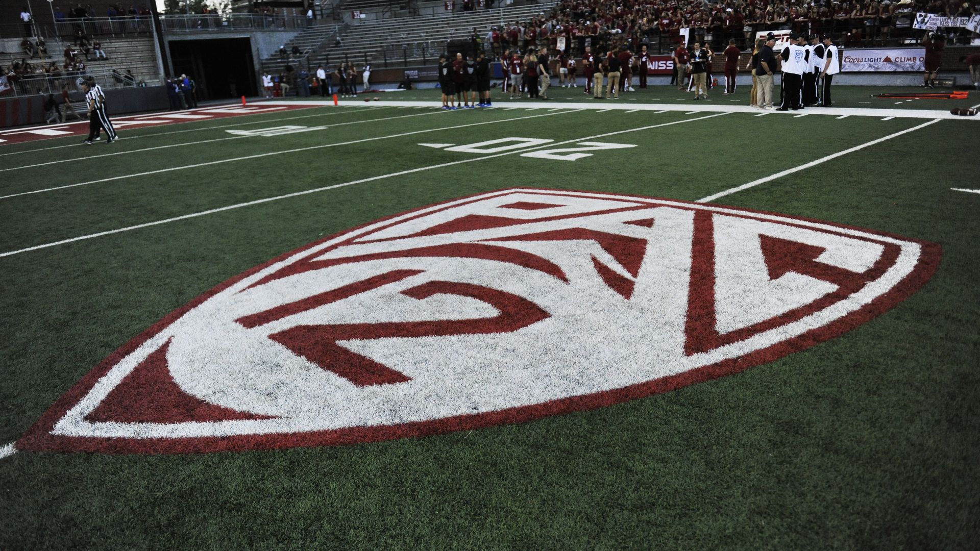 A maroon and white Pac-12 logo is shown on a green football field with spectators in the background in the stands.