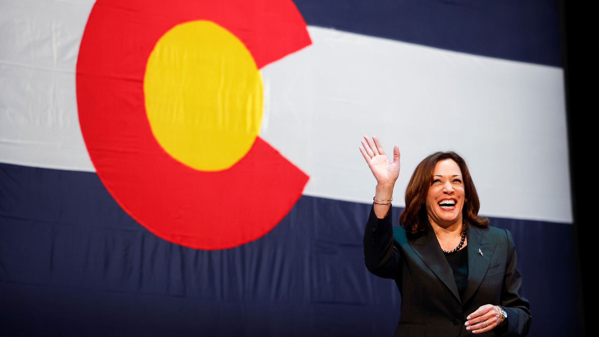 A woman in a suit waves as a large Colorado flag hangs behind her. 
