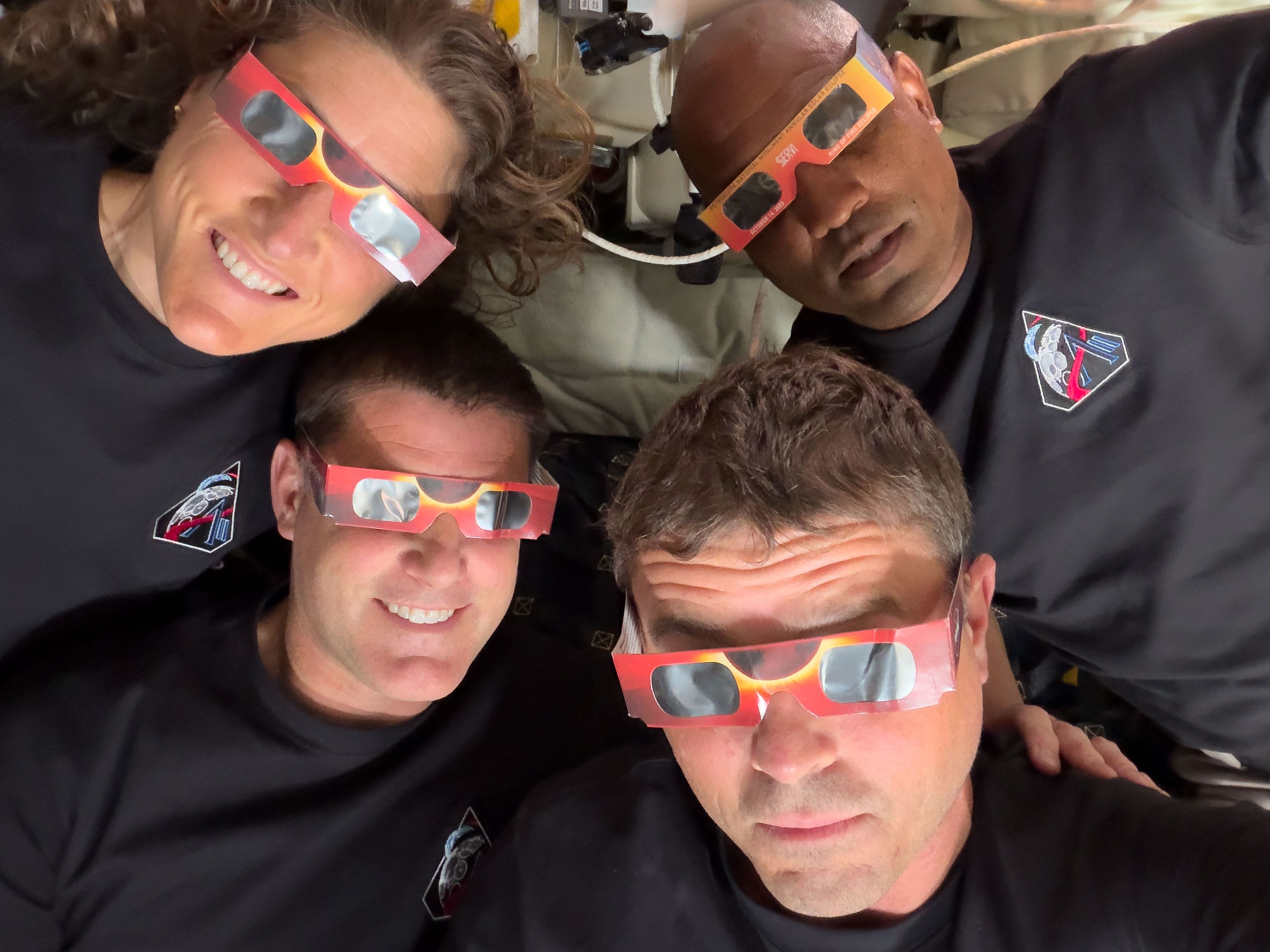 Artemis II's four astronauts wearing in black shirts with an Artemis patch pose for a selfie, wearing red eclipse glasses with gray lenses, smiling, heads tilted toward the camera in the tight Orion crew area