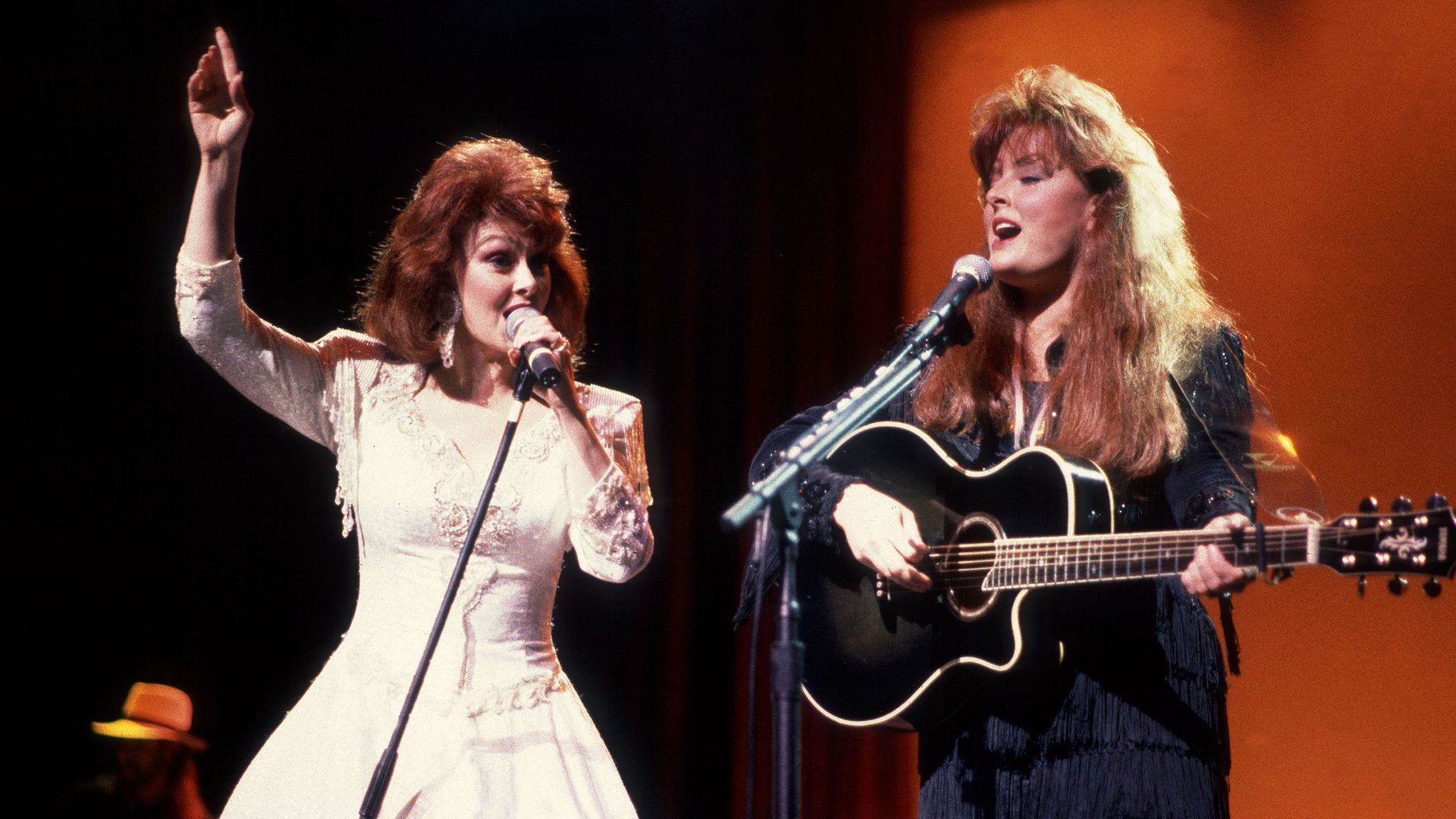 Naomi and Wynnona Judd, with Naomi on the microphone in a white dress and Wynnona holding a black guitar