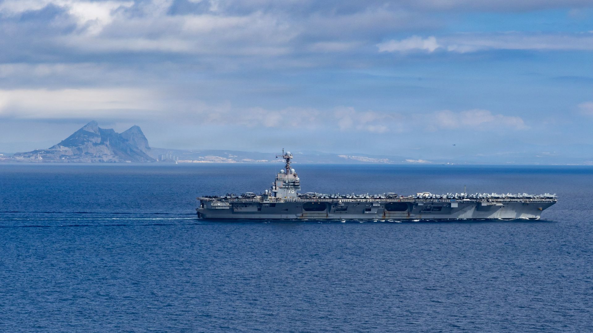 Gray aircraft carrier sailing on blue ocean under partly cloudy sky with rocky mountainous coastline in background.