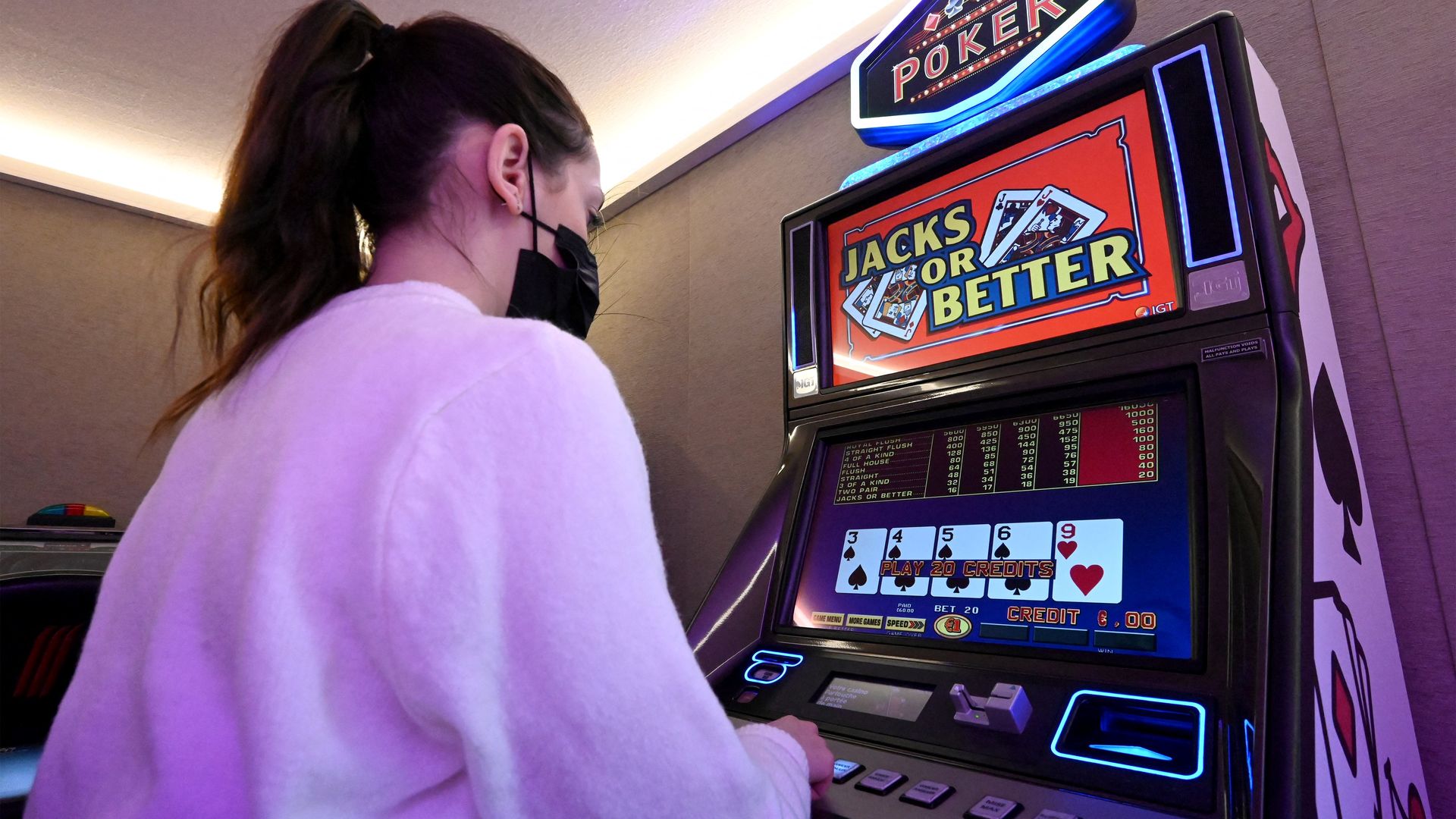 A woman wearing a black mask and white sweater plays a video poker machine with a "Jacks or Better" game on the screen showing cards and credits in a dimly lit room.