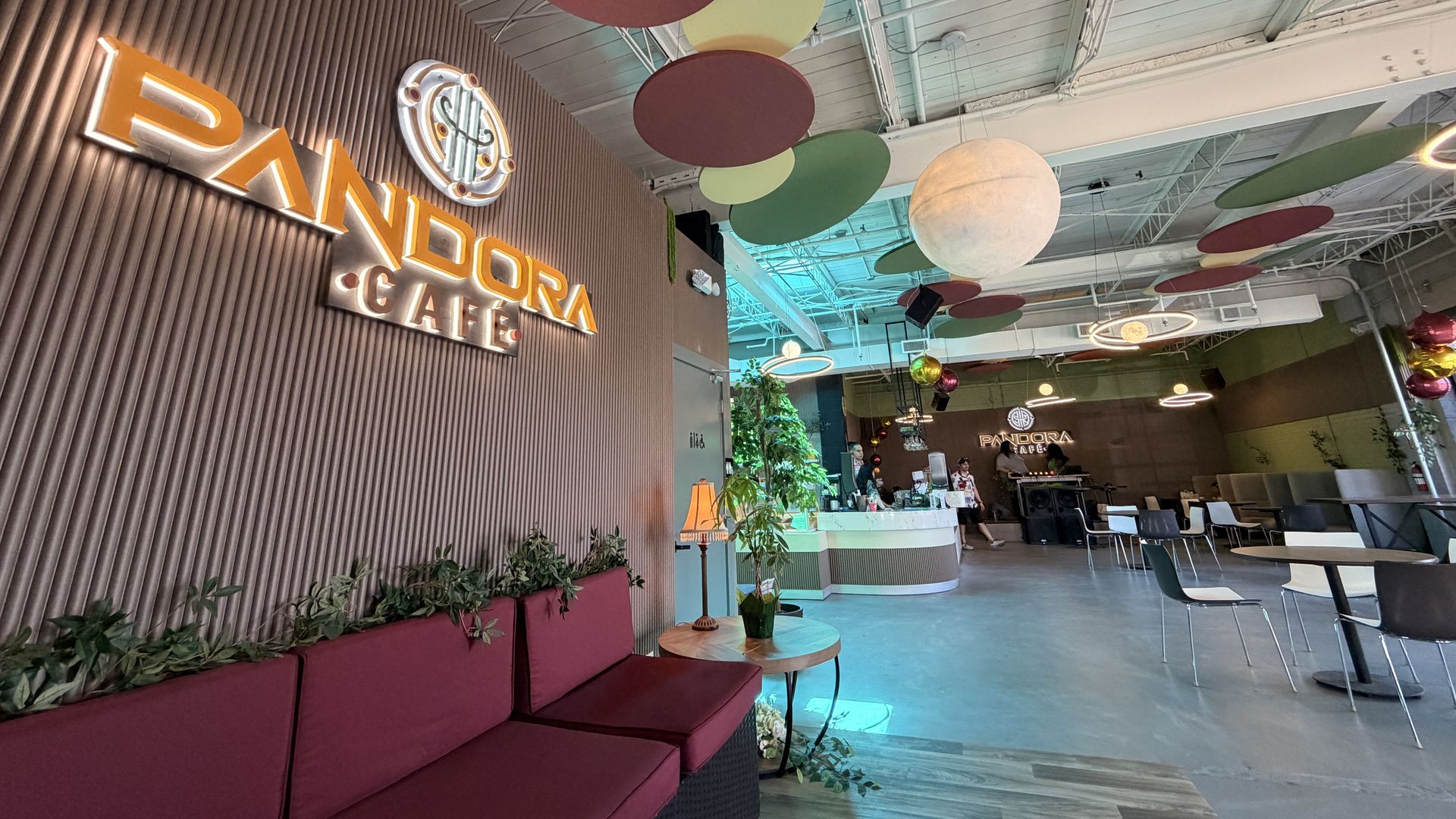 Interior of Pandora Café with a wood-paneled wall and an orange-lit "PANDORA CAFE" sign, colorful hanging circular decorations, red-cushioned couches, plants, and a curved reception desk with patrons.