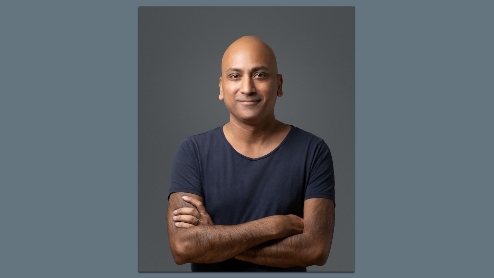Portrait of a bald man in a navy T-shirt with arms crossed, smiling, against a gray gradient backdrop.