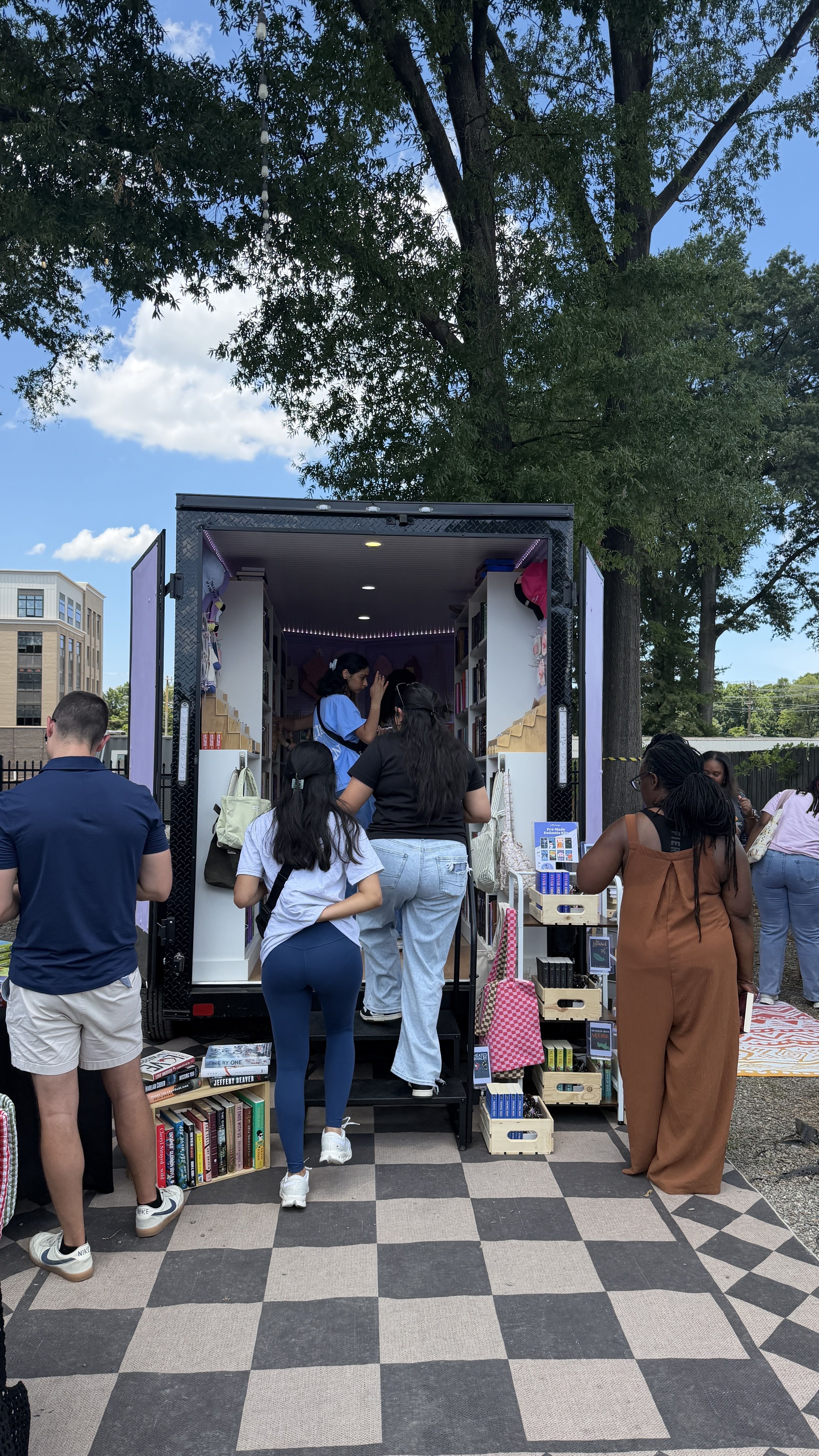 People browse a purple open-back mobile bookstore truck parked outdoors; shelves and books inside, crates of books outside, under a leafy tree on a sunny day with a checkered pavement.