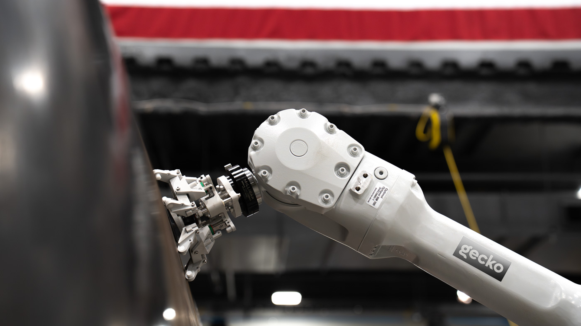 White robotic arm labeled gecko grips a bolted metal housing with a circular cover, in a workshop with a red stripe and a yellow cable in the background.