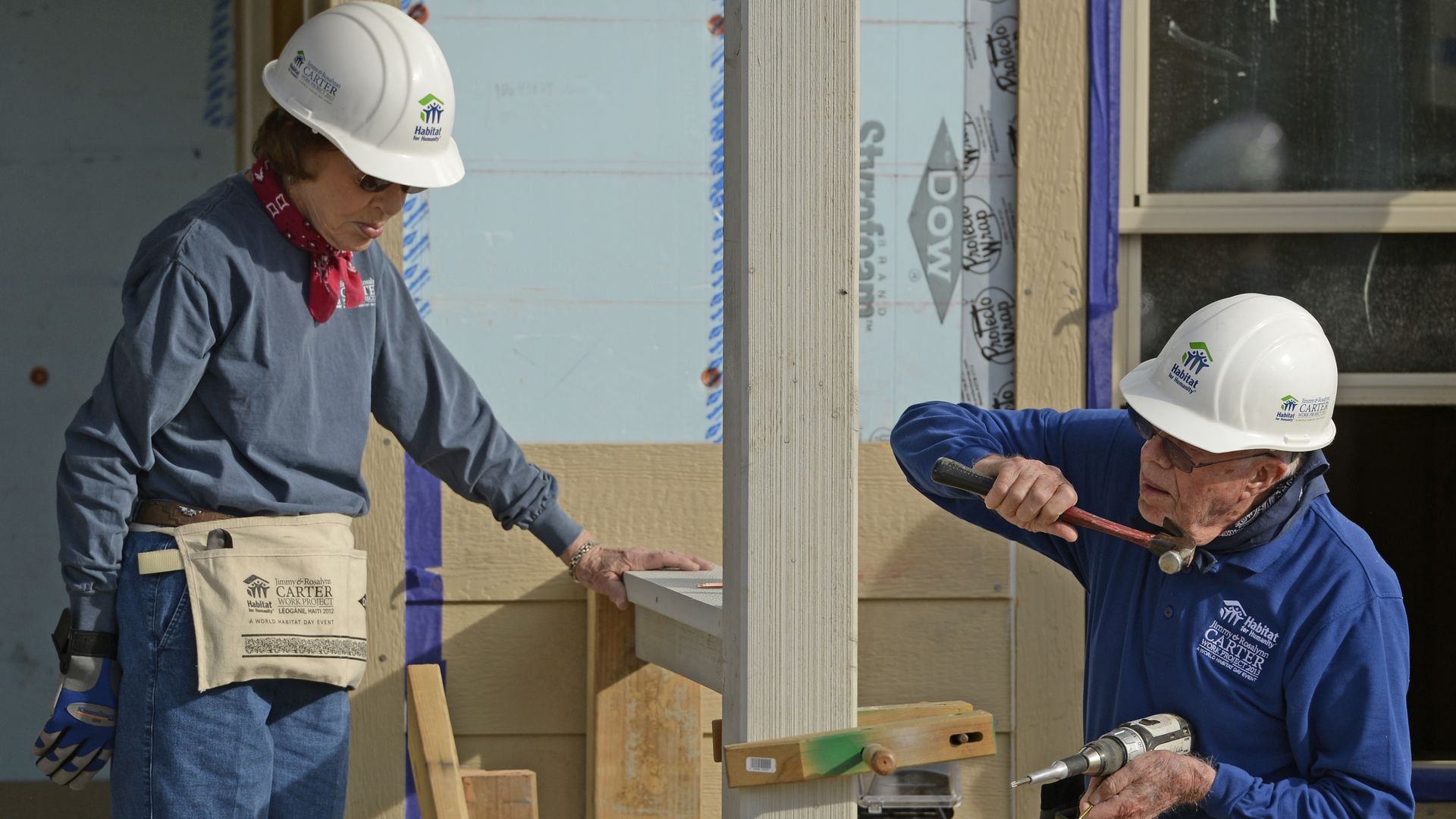 Two people wearing white hard-hats and construction gear help construct a home. A man wields a hammer in his right hand and holds a drill in his left hand as a woman watches.