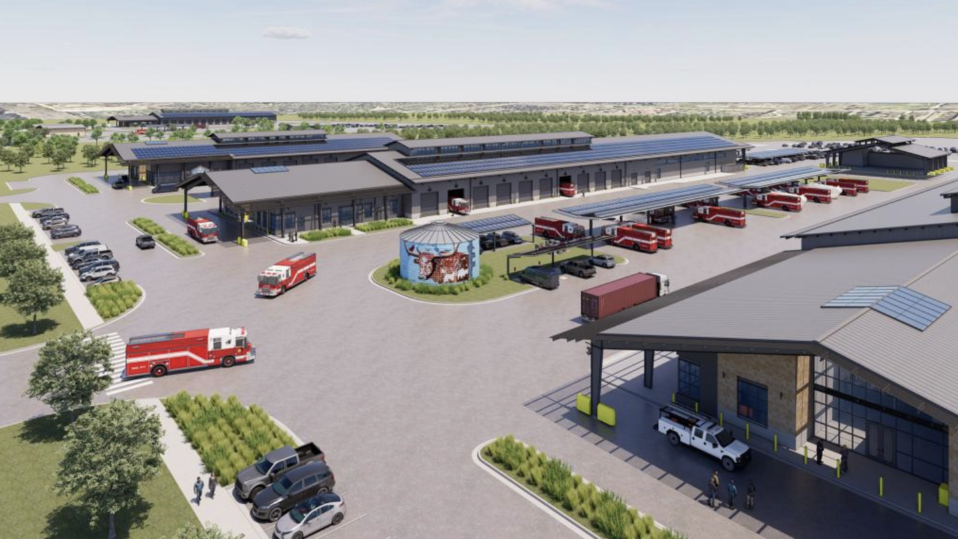 Aerial view of a fire station complex with multiple red fire trucks, gray buildings with solar panels, parked cars, a blue cylindrical structure with a mural, and people walking nearby on a clear day.
