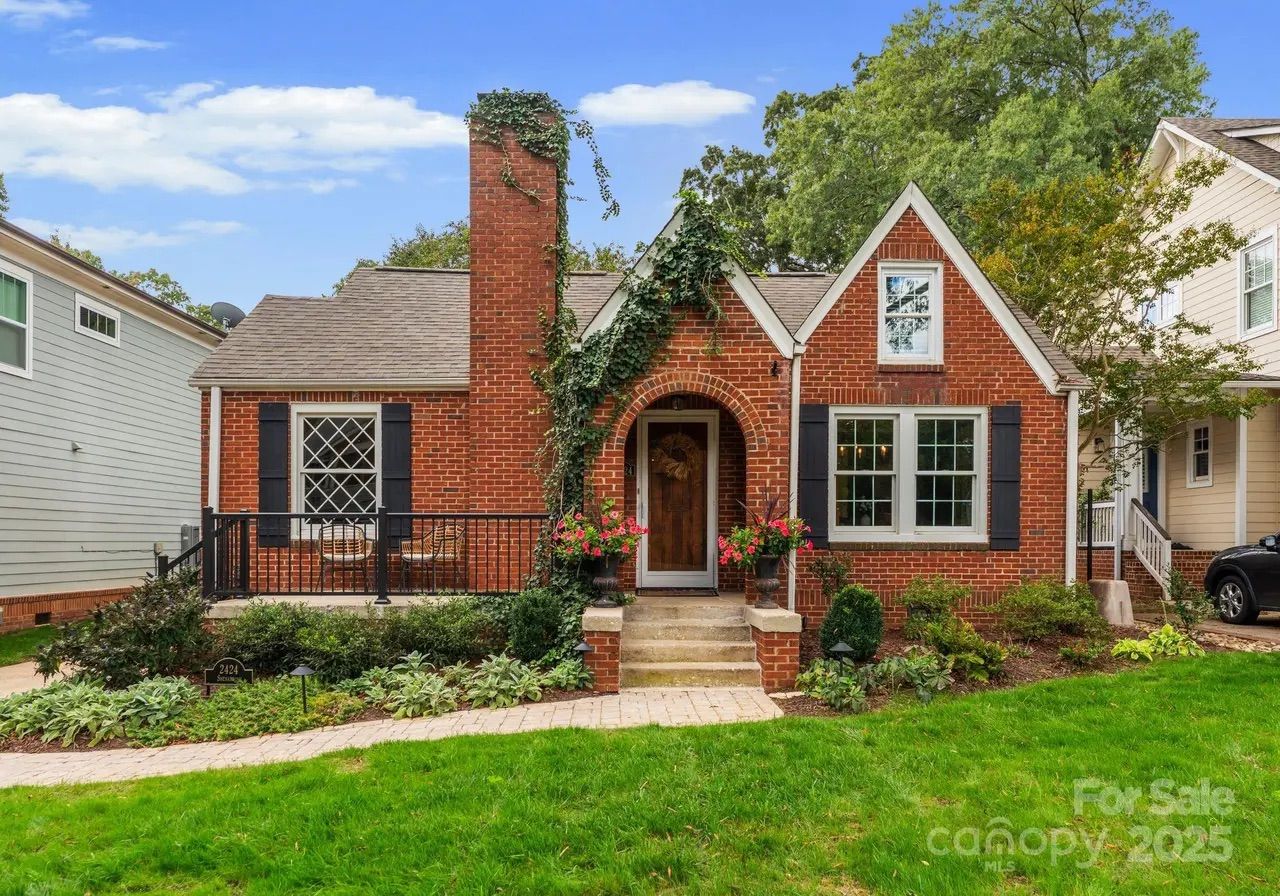 Brick house with black shutters, arched doorway, ivy on chimney, flower pots at entrance, green lawn, and blue sky with clouds, neighboring houses and trees visible.