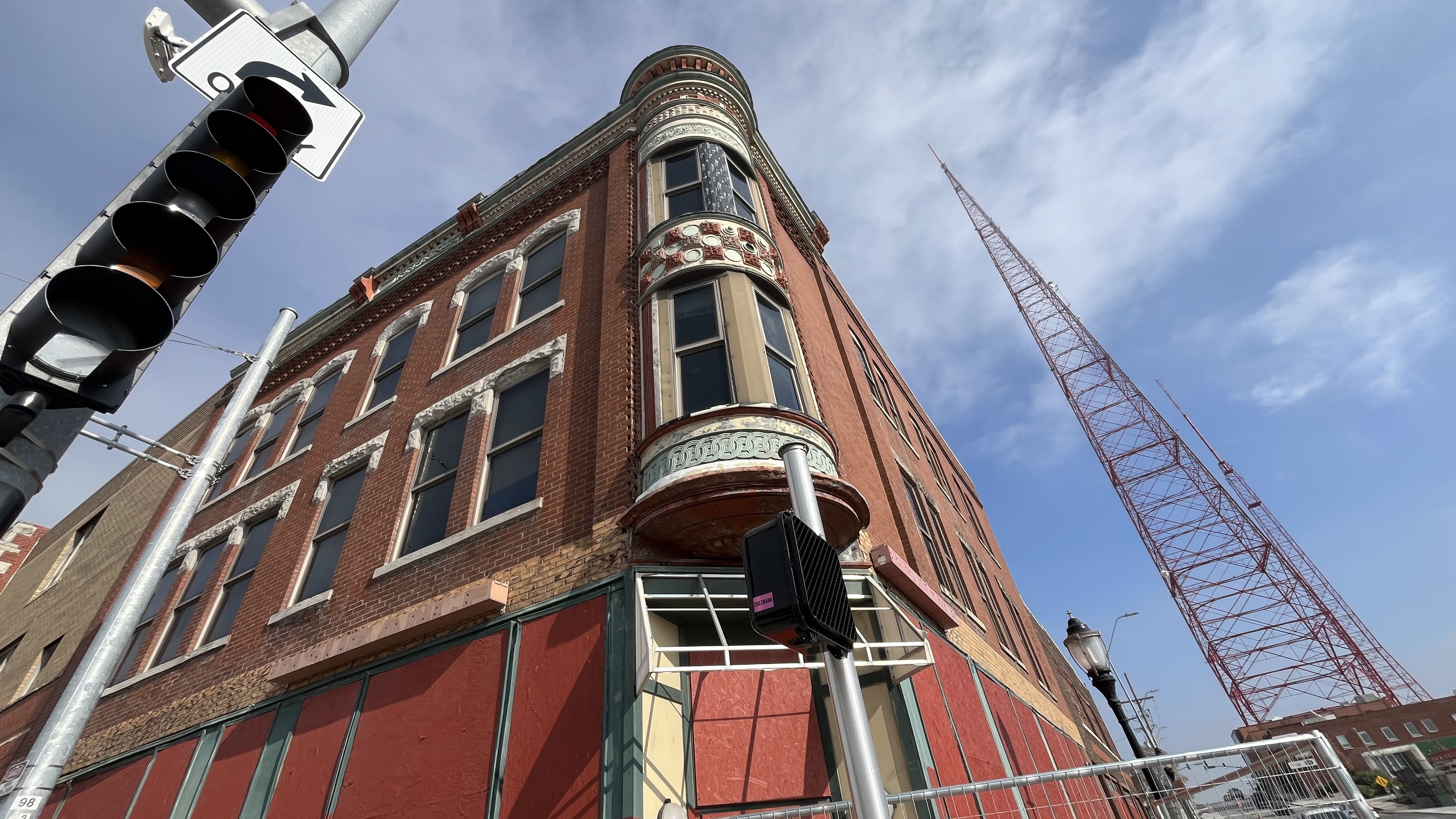 Red brick corner building with ornate rounded bay window and boarded-up lower windows, adjacent to a traffic light and tall red radio tower under a partly cloudy blue sky.