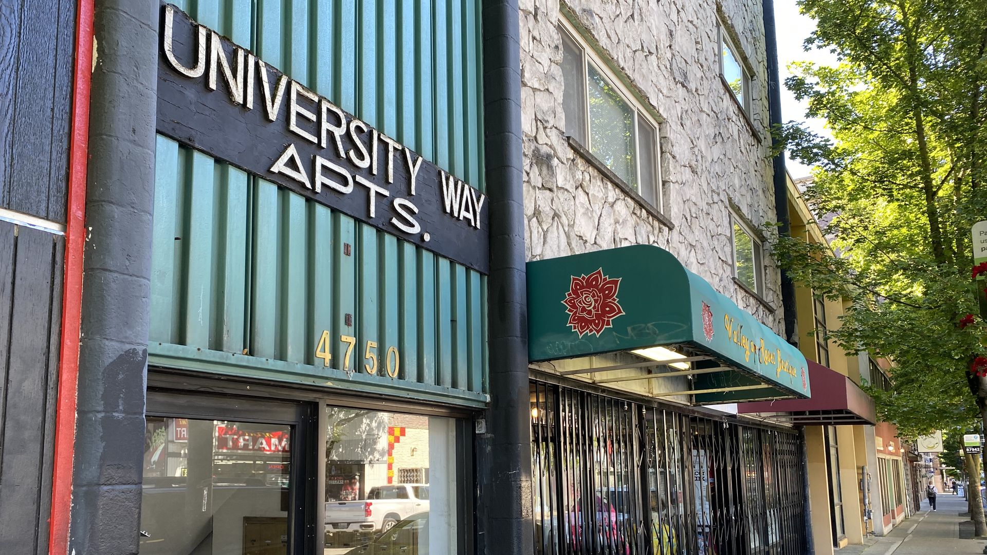Building with text that says "University Way Apts" and a store awning behind it, along a sidewalk. 