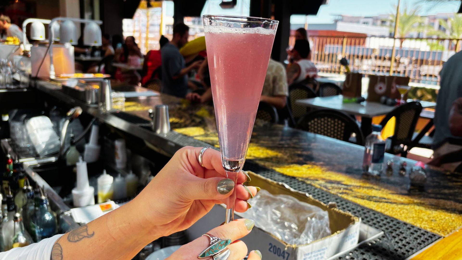 Foreground pink cocktail in a tall champagne flute held by a tattooed hand; a busy bar scene with blurred customers, open-air seating, and palm trees in the background.