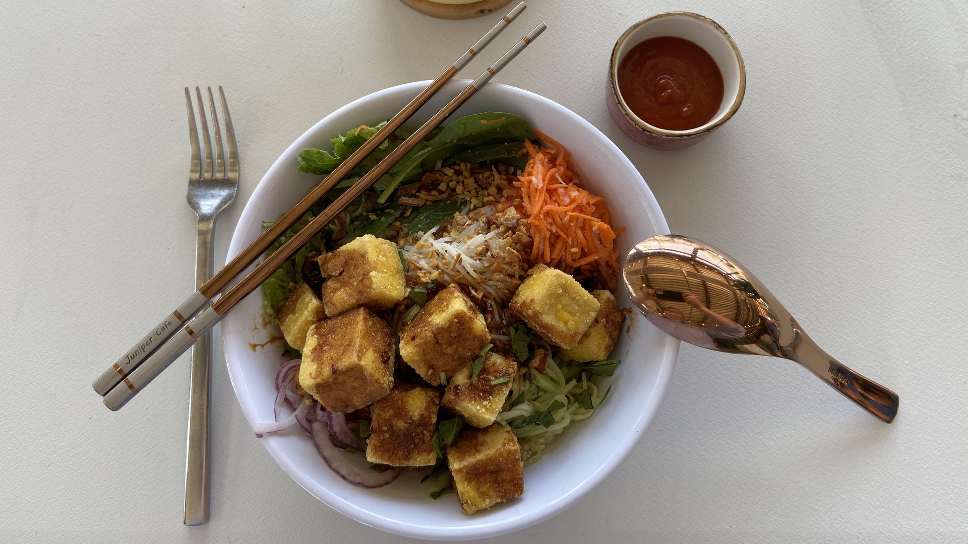 A photo of a noodle salad bowl with fried tofu, greens and carrots, framed by gold chopsticks and a soup spoon and a small cup of Sriracha