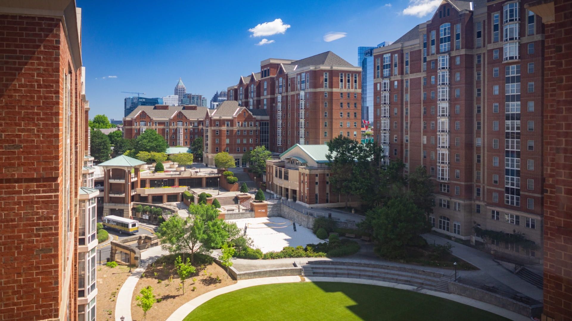 Wide campus courtyard bordered by red-brick dorms and glass towers, with a green lawn, curved paths, trees, and a bright blue sky.