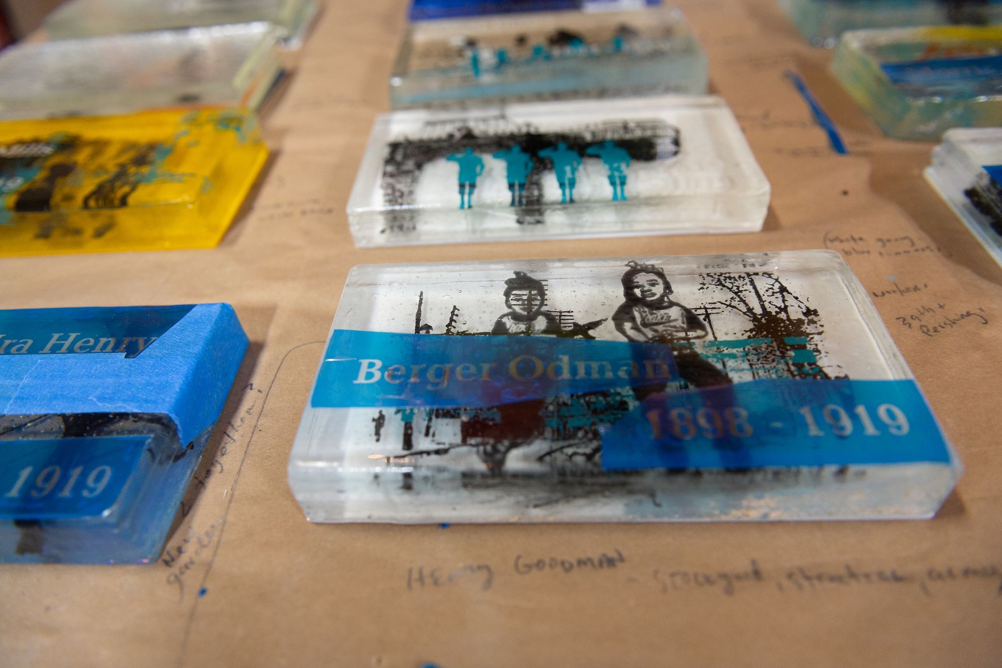 On a tan table, several small art boxes or books arranged in rows. Foreground shows a white box with a black-and-teal illustration of two silhouetted people and a blue label; surrounding are yellow and blue covers.