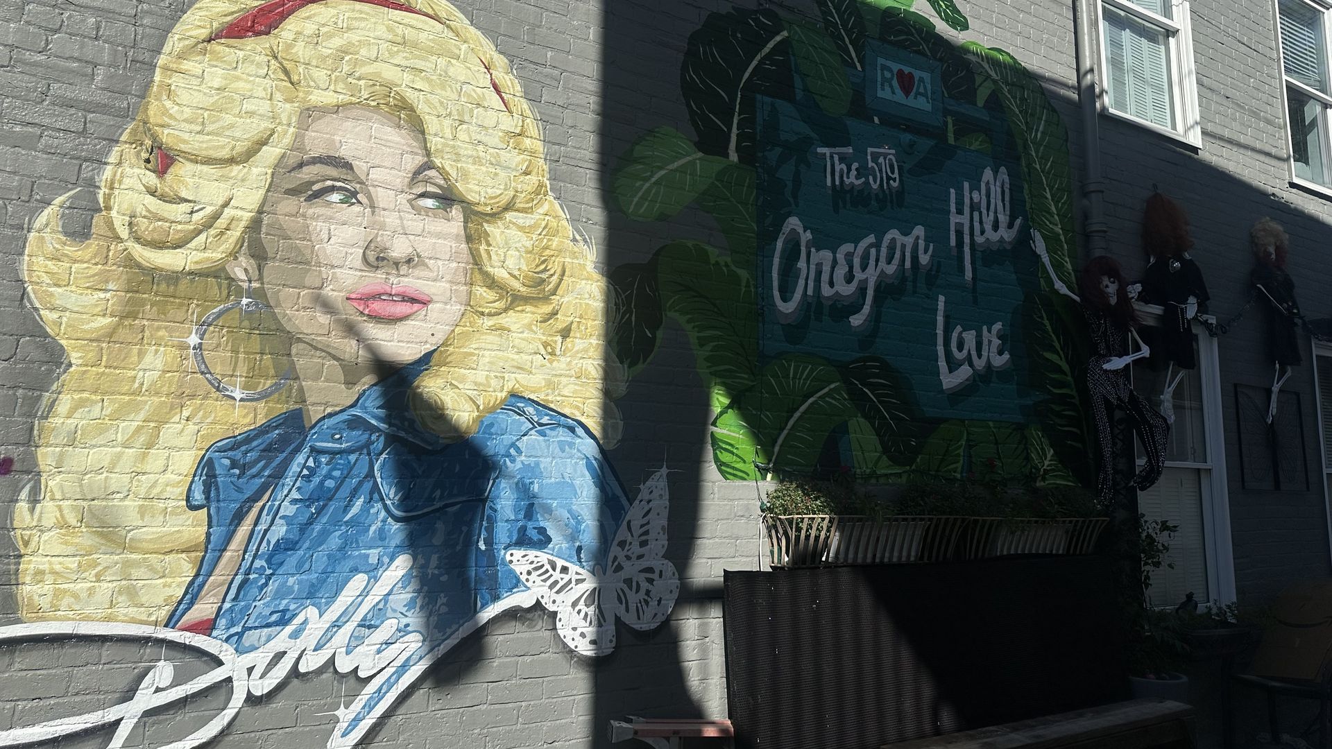 Large mural on a gray brick wall shows a blonde woman in a blue shirt, with big green tropical leaves and a blue panel reading "The 519" and a heart symbol; window frames and shadows above.