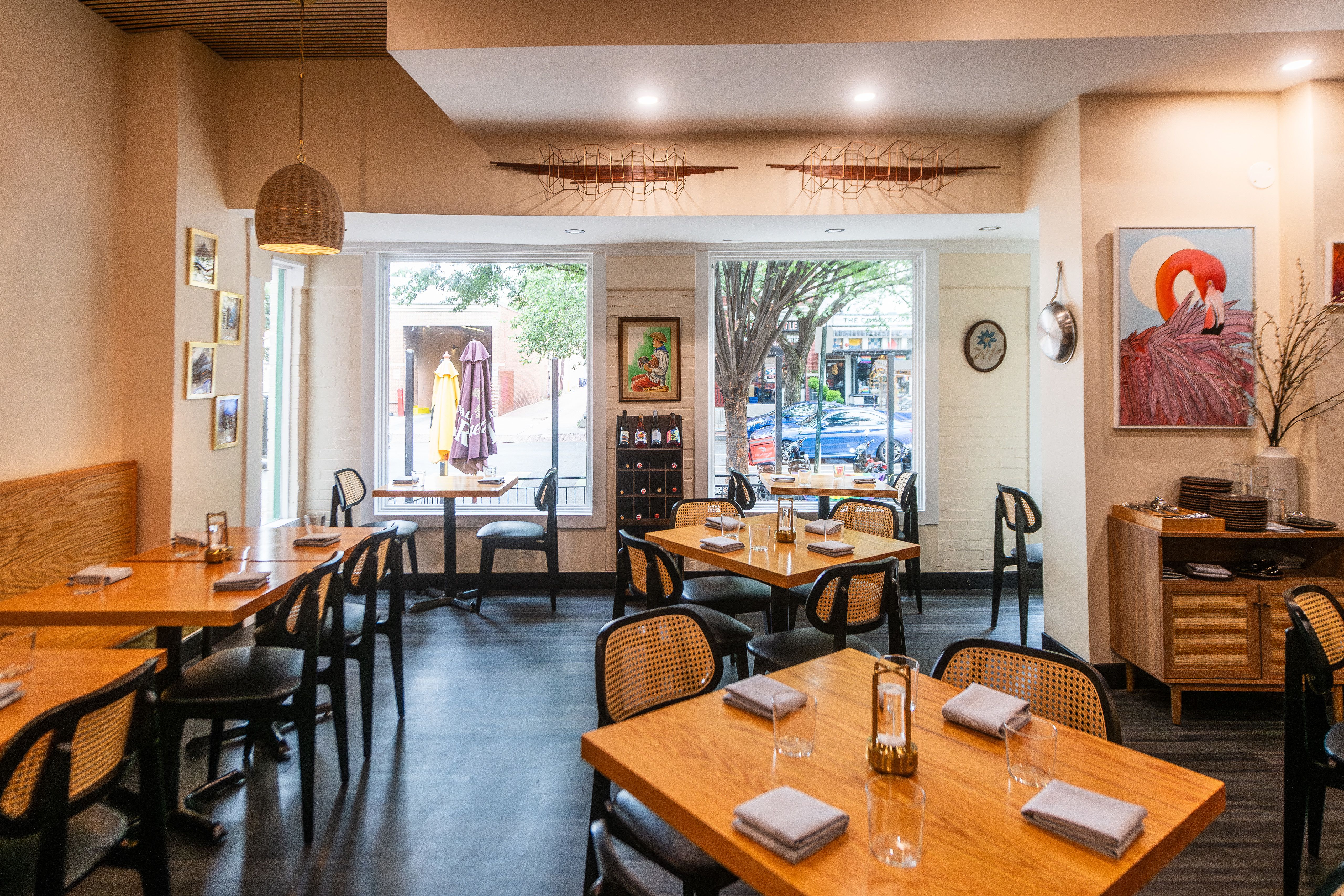 Bright restaurant interior with wooden tables, black chairs with cane backs, white walls, hanging lamp, framed artworks including a pink flamingo, and large windows showing street outside.