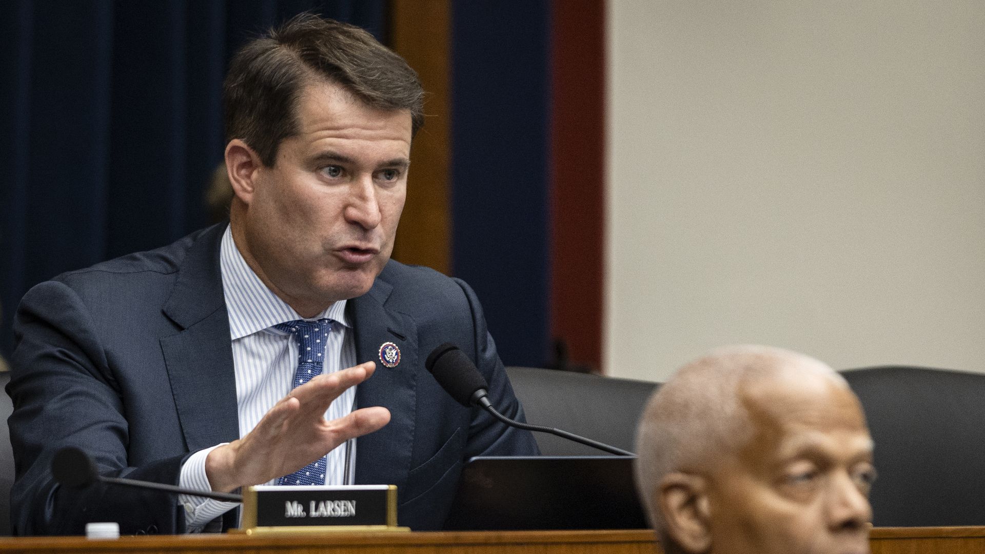 Rep. Seth Moulton, wearing a gray suit and raising his hand at a wooden committee dais in front of a blue curtain and a white wall.